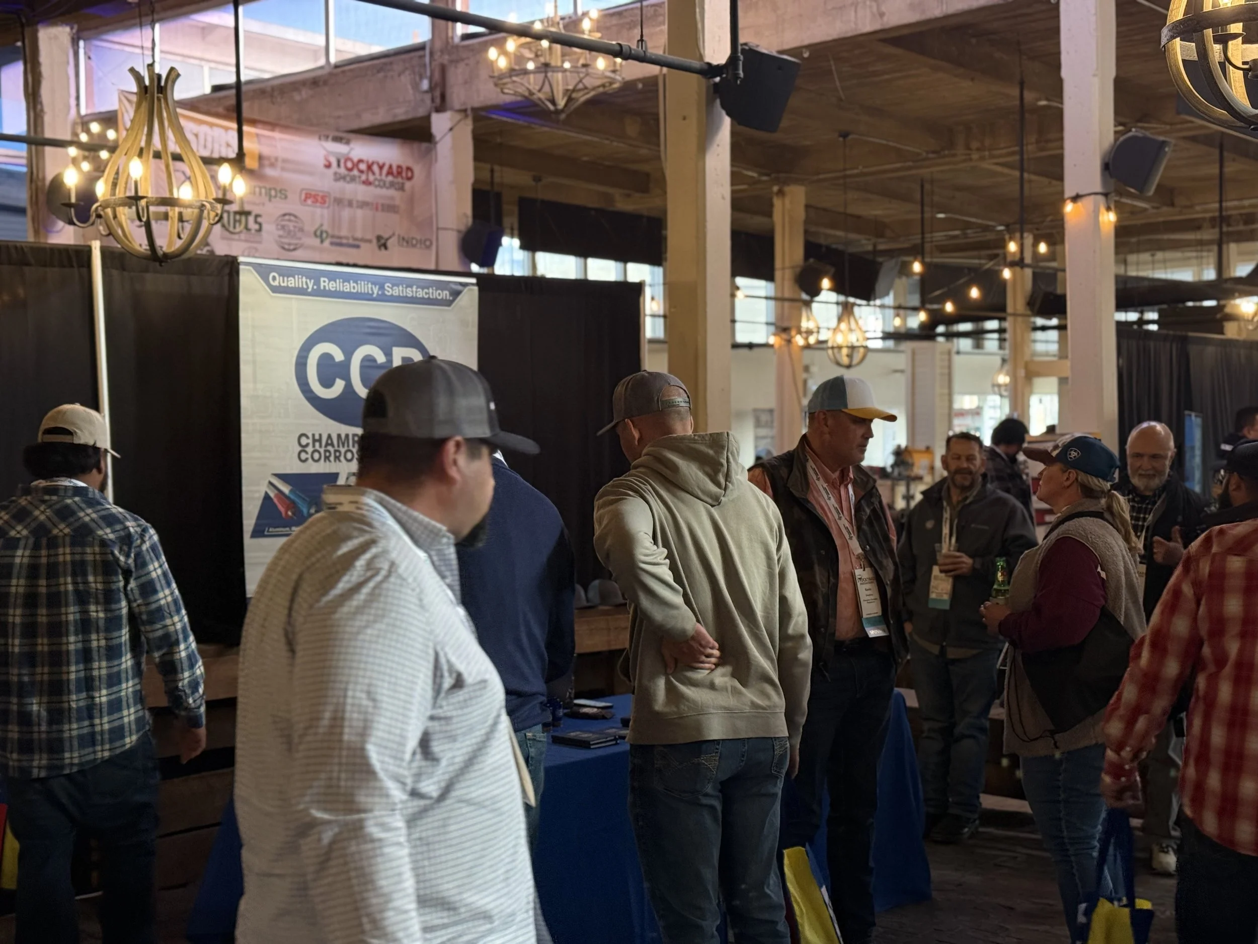 People attending an indoor trade show or conference, standing around booths with banners. One prominent banner reads "Championship Corrosion" with a logo and slogan about quality and reliability. The venue features wooden beams, warm lighting, and ch