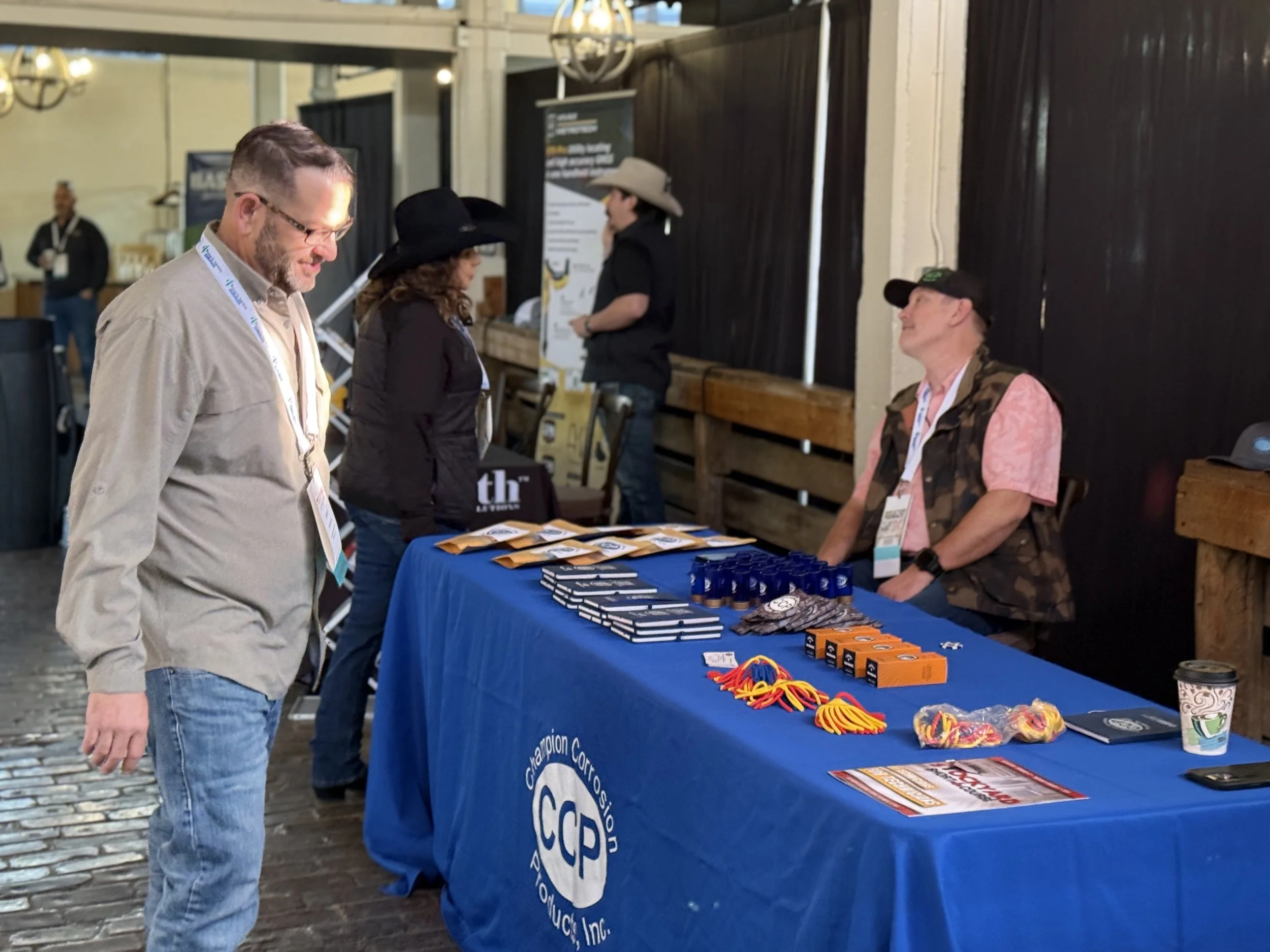 A man in a beige shirt and jeans is looking at a booth table with CCCP logo, man and woman manning it at an event.