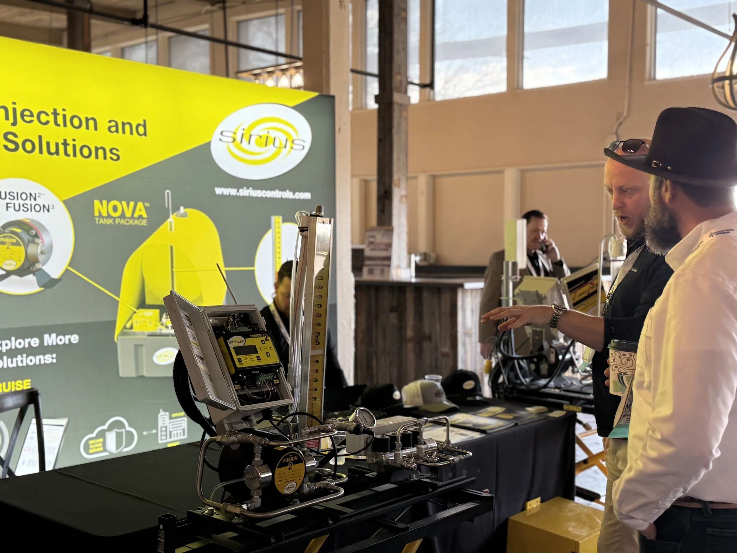Two men talking at a trade show booth with a large yellow and green display about Sirius controls and injection solutions. The booth features equipment and explanation boards.