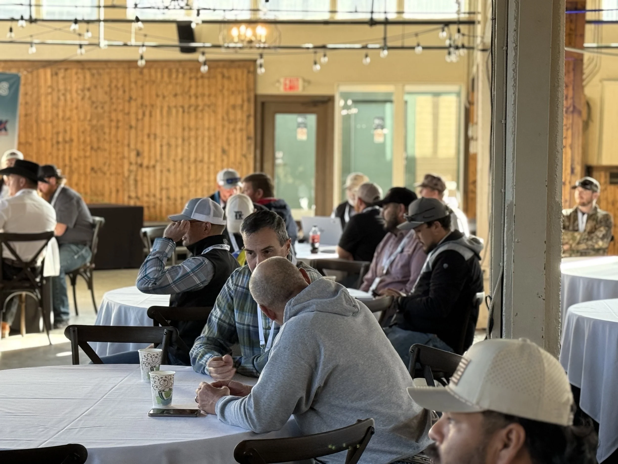 Indoor gathering of men seated at round tables with white tablecloths, some using phones, wearing casual attire including hats and hoodies, with wooden walls and string lights in the background.