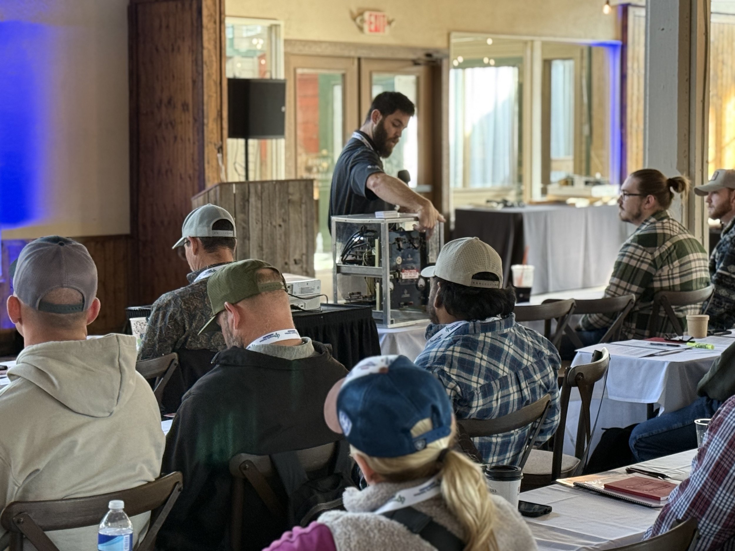 A man demonstrating a mechanical or technical device to an audience in a conference room with wooden decor and large windows.