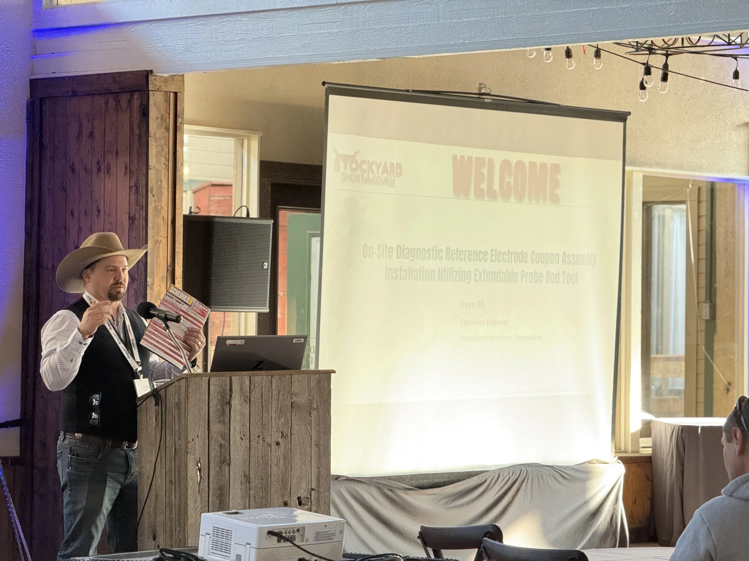 A man wearing a cowboy hat and vest is standing at a wooden podium, holding a pamphlet and speaking into a microphone during a presentation in a rustic, wood-paneled room. There is a large screen displaying a welcome slide in the background.