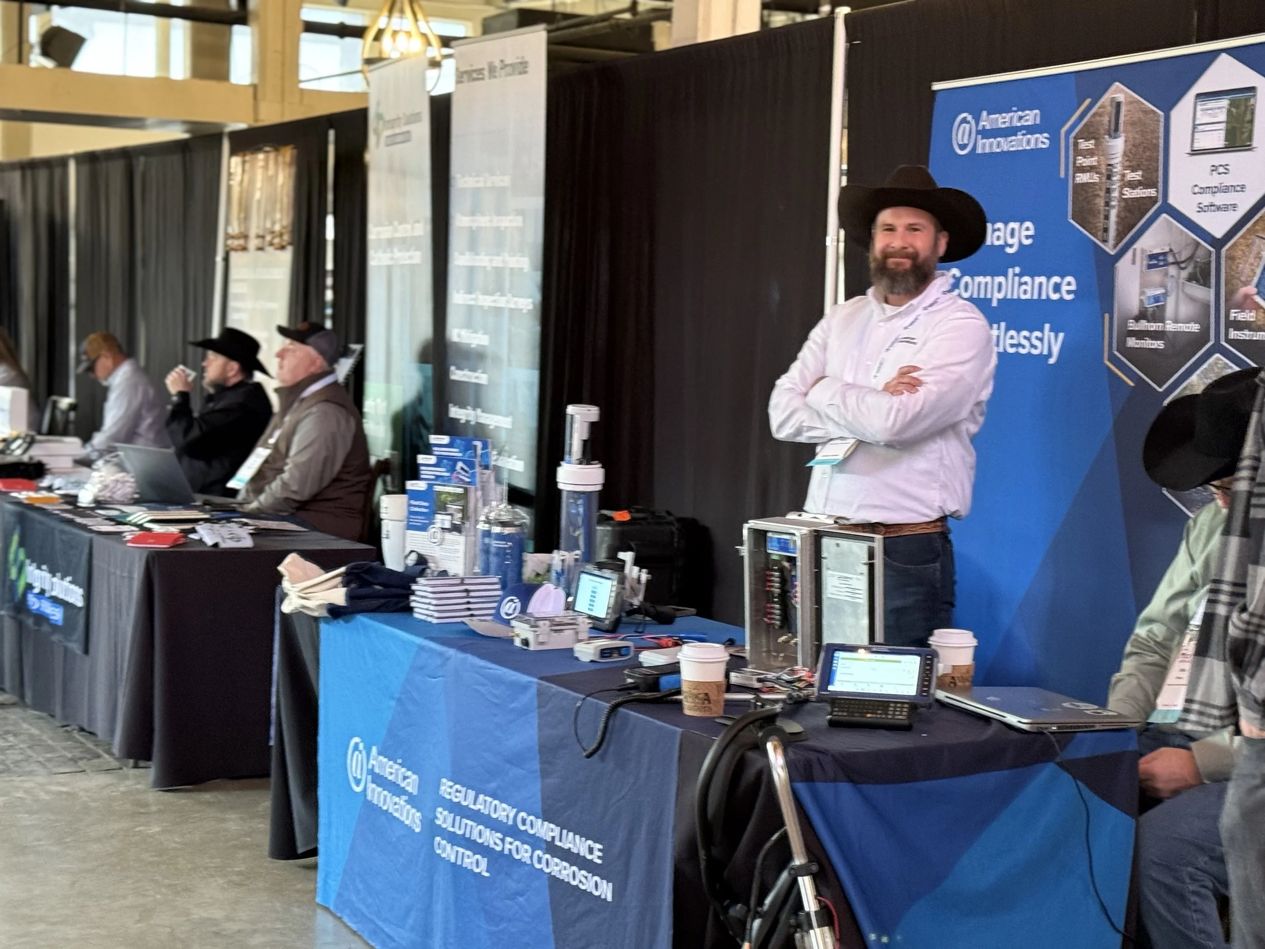 Man with beard wearing a black cowboy hat and white shirt standing at a trade show booth, smiling with arms crossed. The booth has a blue banner with the American Innovations logo and displays regulatory compliance solutions.
