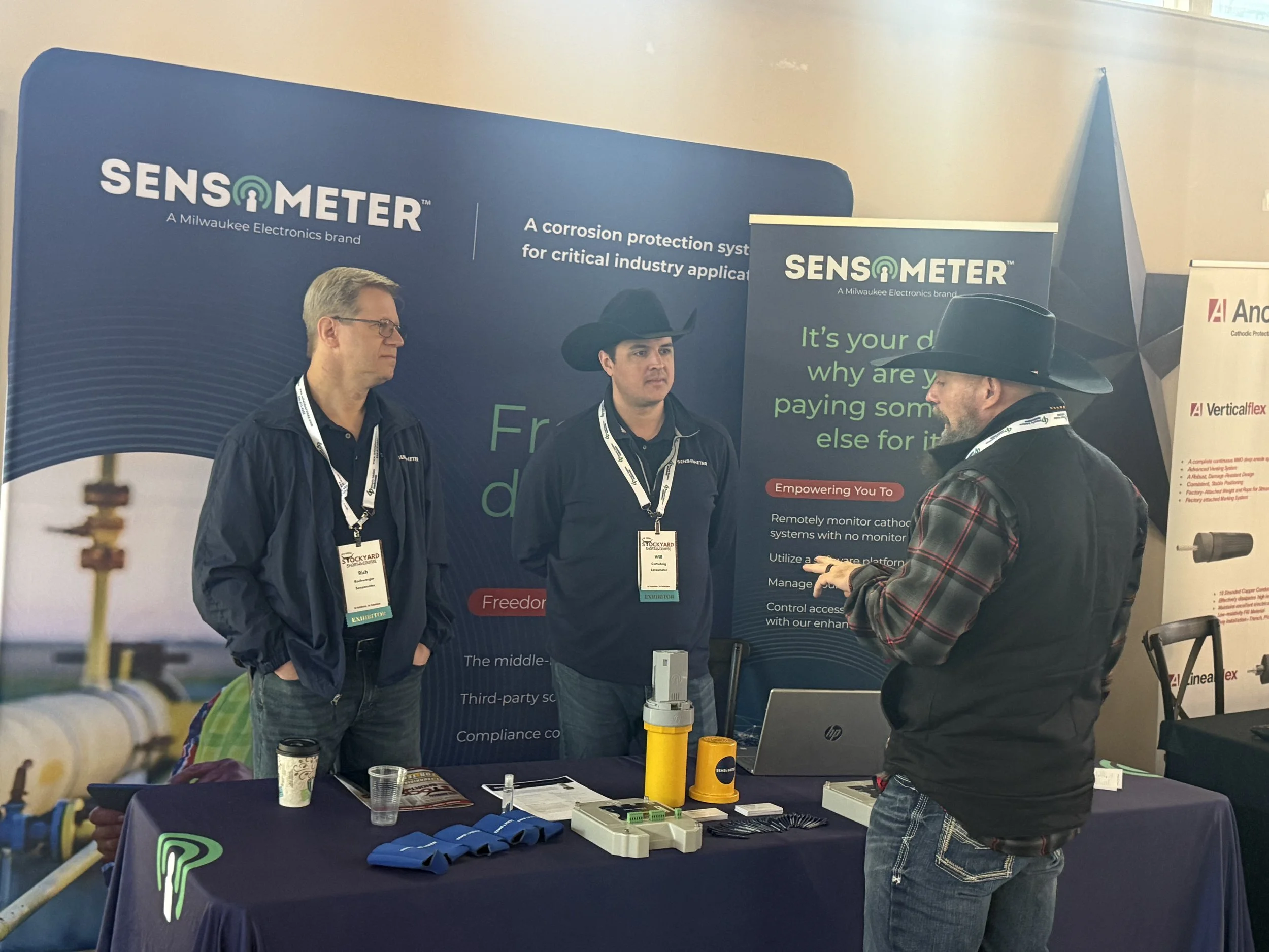 Three men stand at a trade show booth for Sensometer, a Milwaukee Electronics brand, discussing products. Two men are behind the booth, wearing dark jackets and lanyards, while the third man, wearing a cowboy hat and vest, is speaking with them. The 