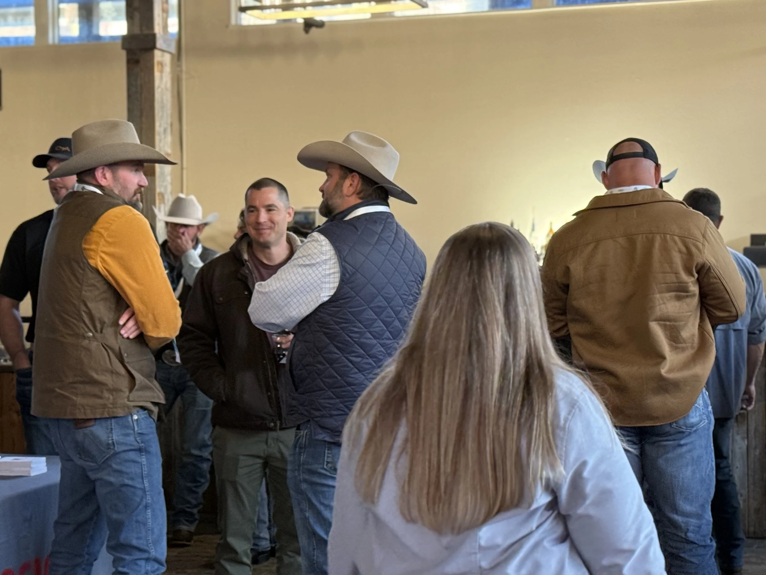 Group of people at a social gathering, some wearing cowboy hats and casual attire, talking and smiling in an indoor setting.