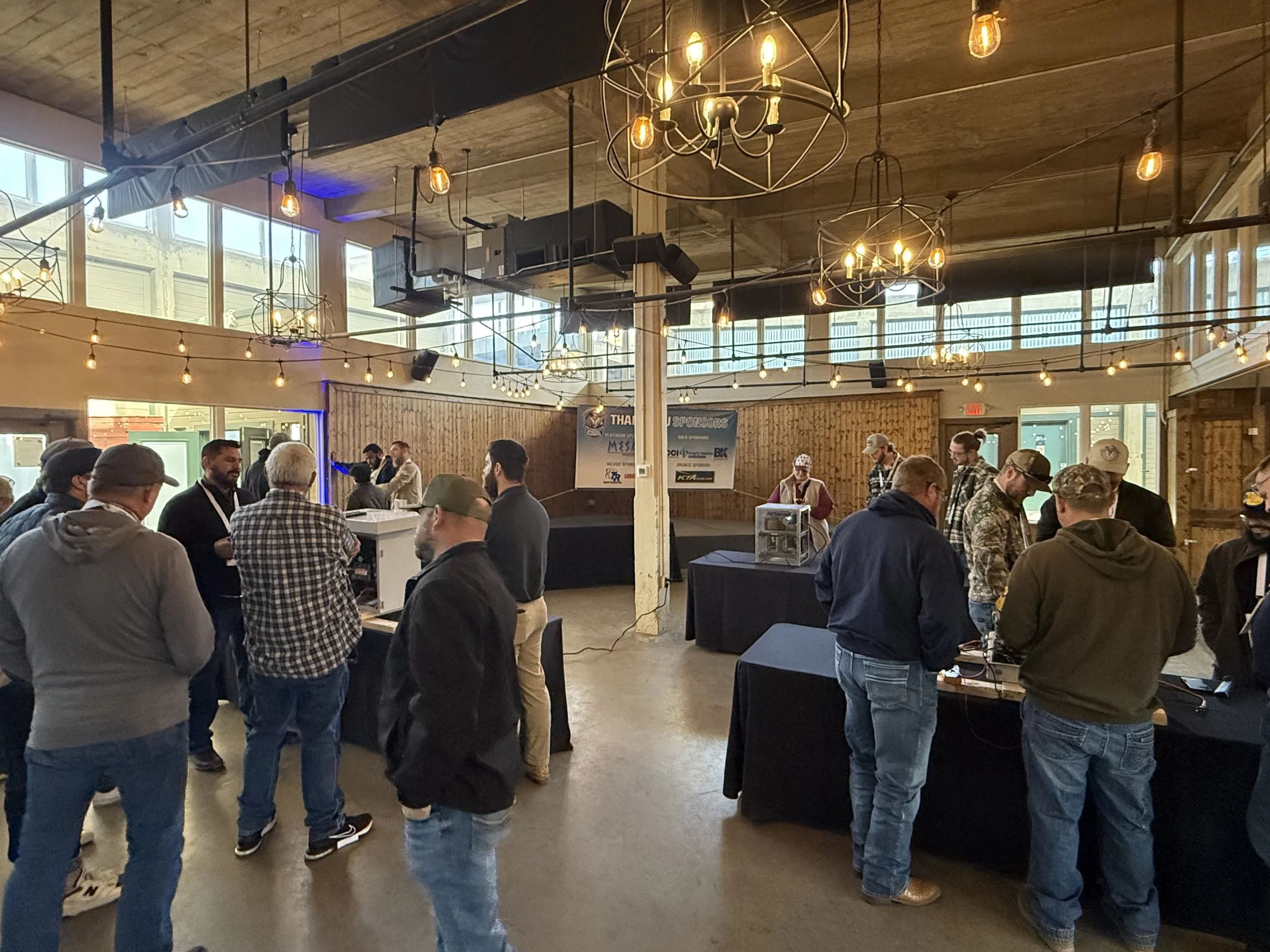 People gathered in a large indoor space with high ceilings, wooden walls, and hanging string lights and chandeliers, attending an event with registration tables and a stage area.