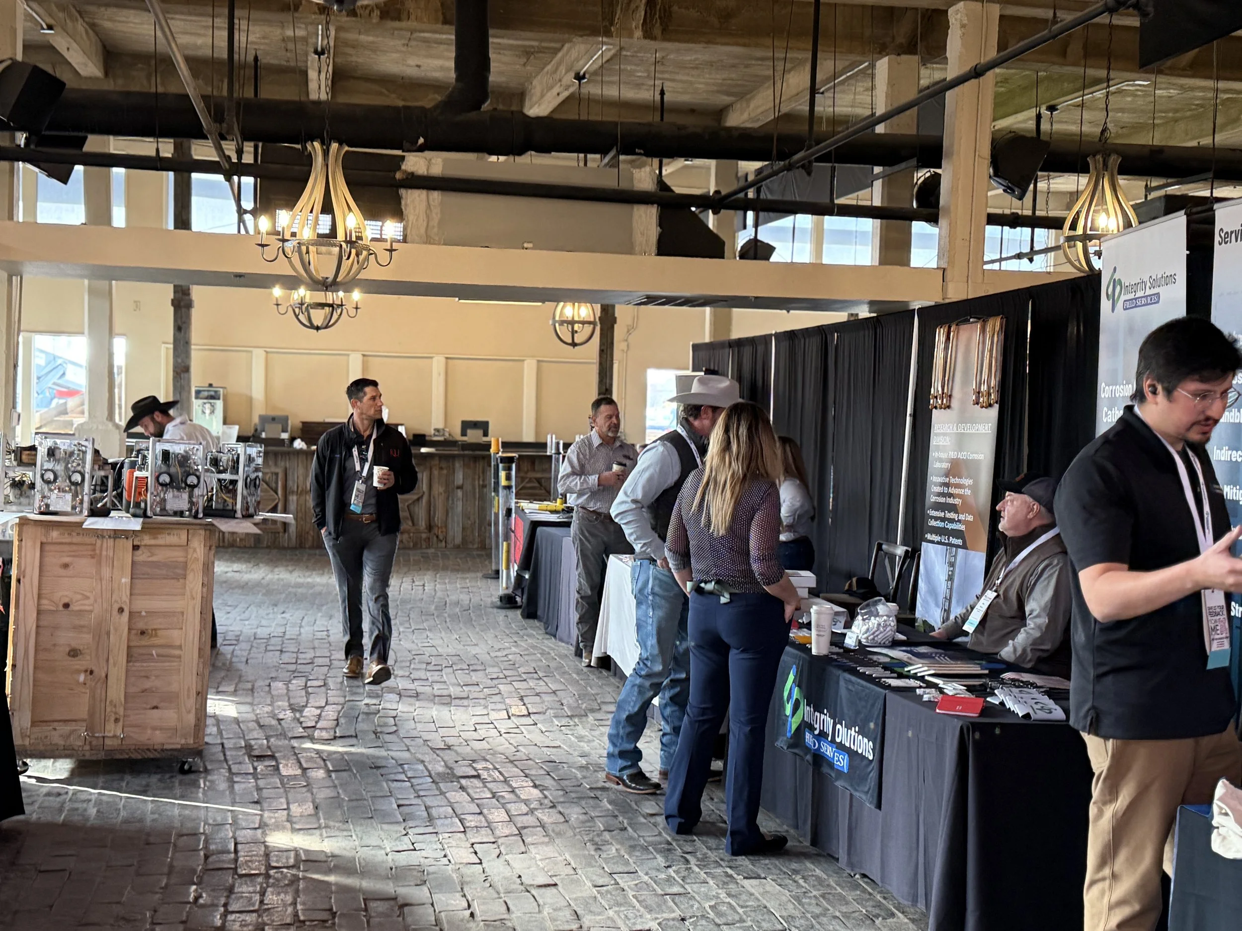 People attending a convention or trade show in a spacious, industrial-style indoor setting with brick flooring, chandeliers, and booths displaying products and informational materials.
