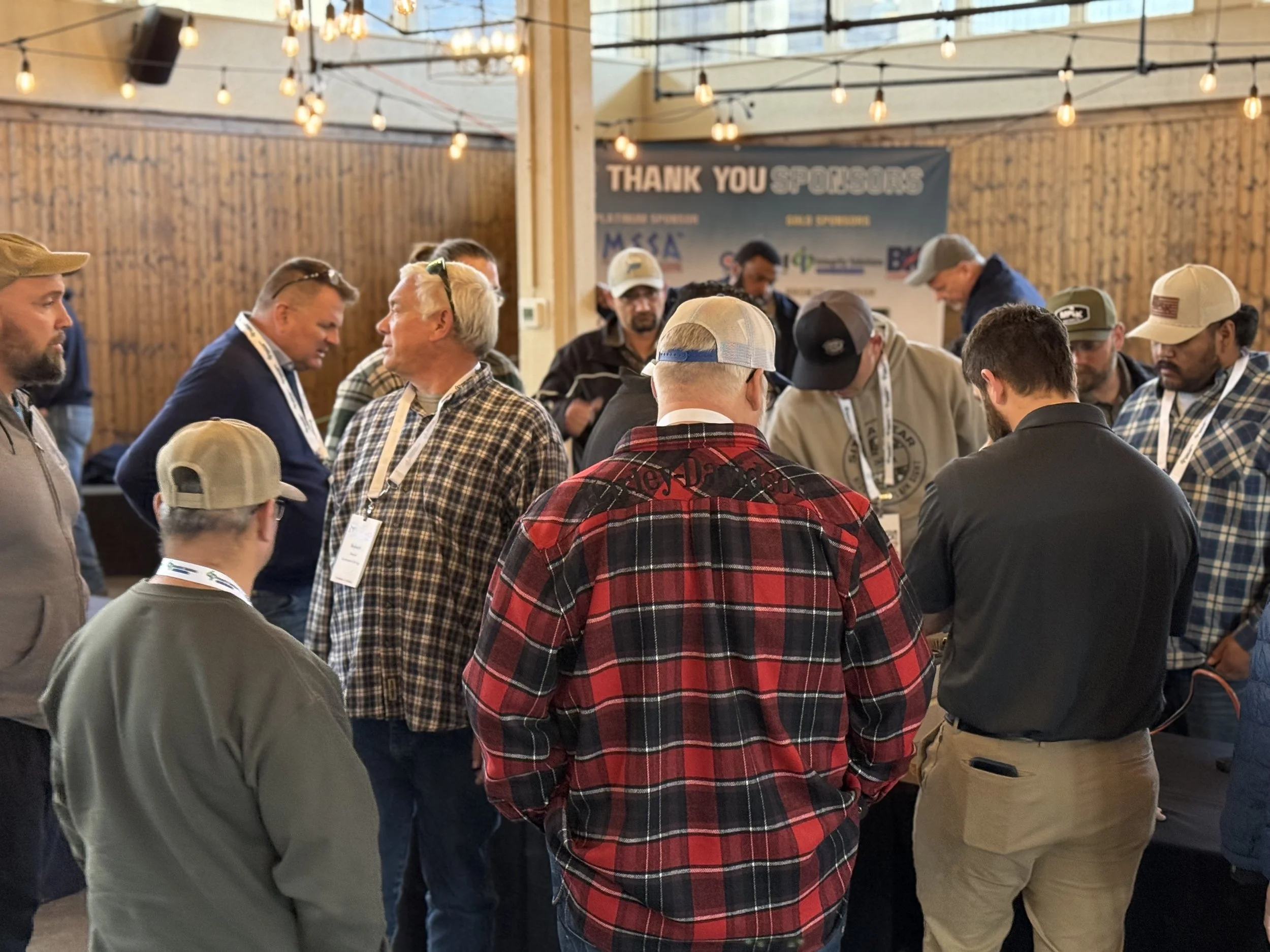 Group of people gathered at an indoor event with a wooden theme, string lights, and a banner thanking sponsors in the background.