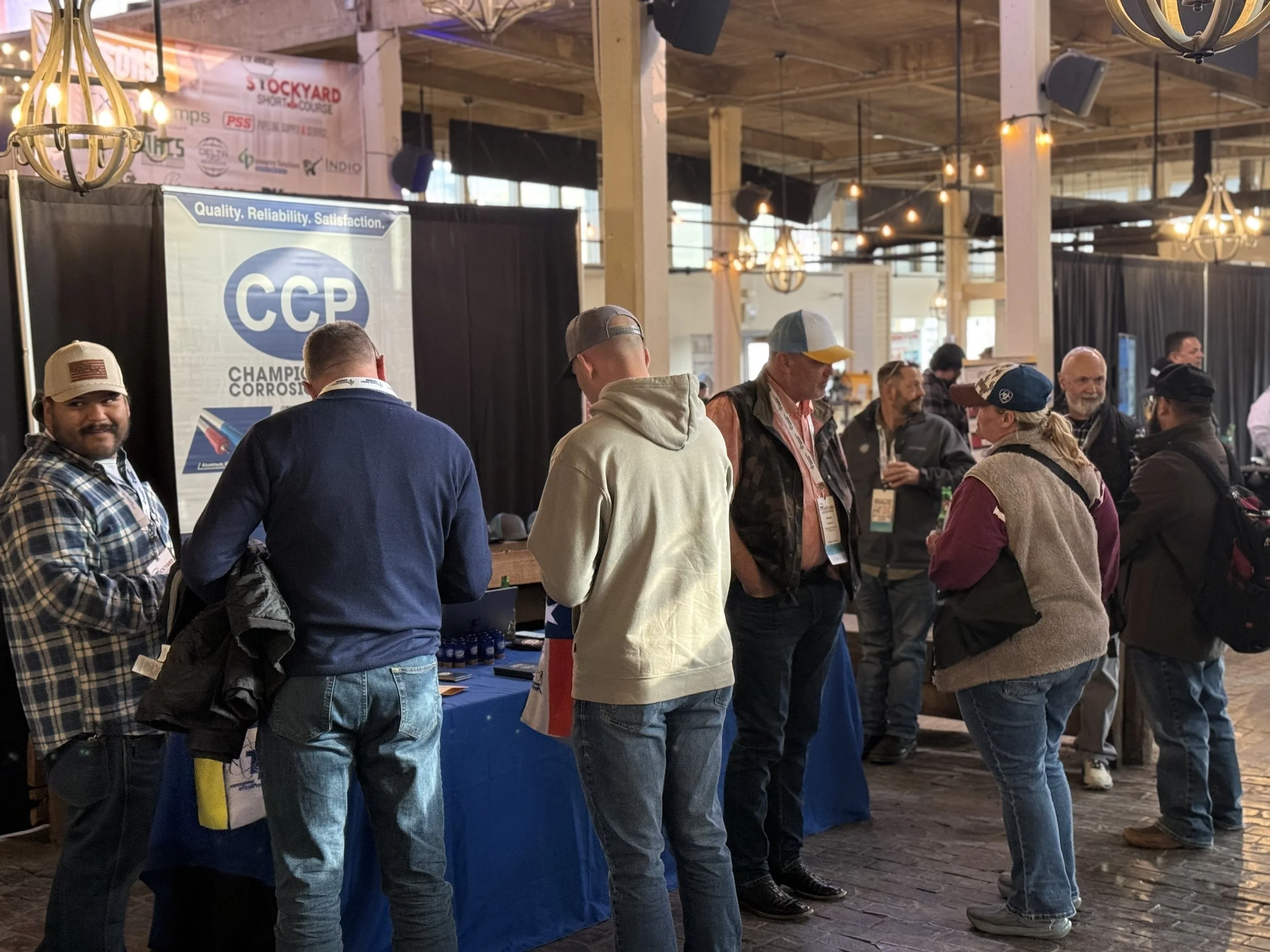 People gather at a trade show booth with a CCP display, engaging with the vendor. The booth has a table with products or information, and the background features banners and hanging lights in a large, open indoor space.