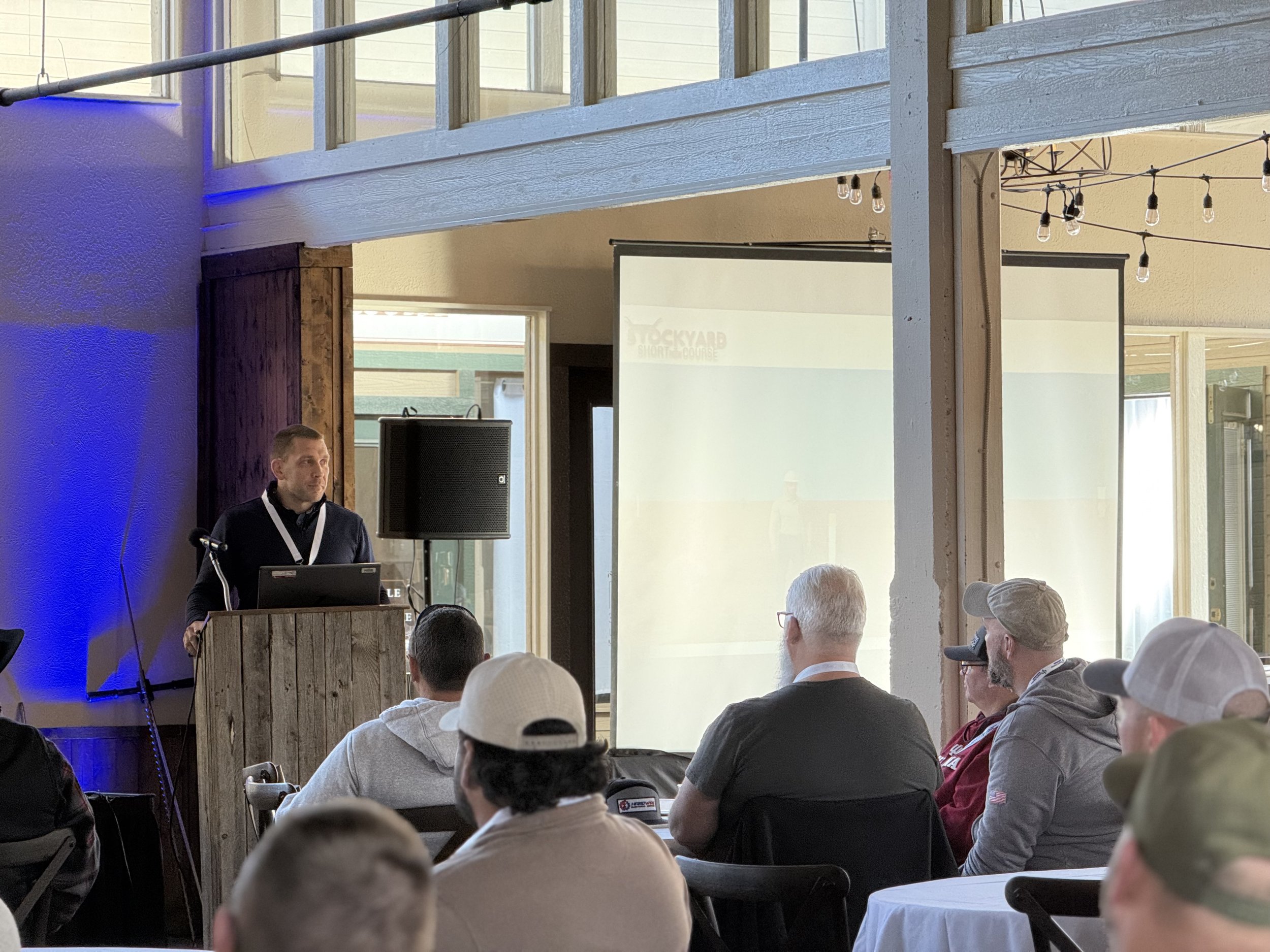 A man stands at a wooden podium giving a presentation to an audience seated in a room with natural and artificial lighting, large windows, and string lights overhead.