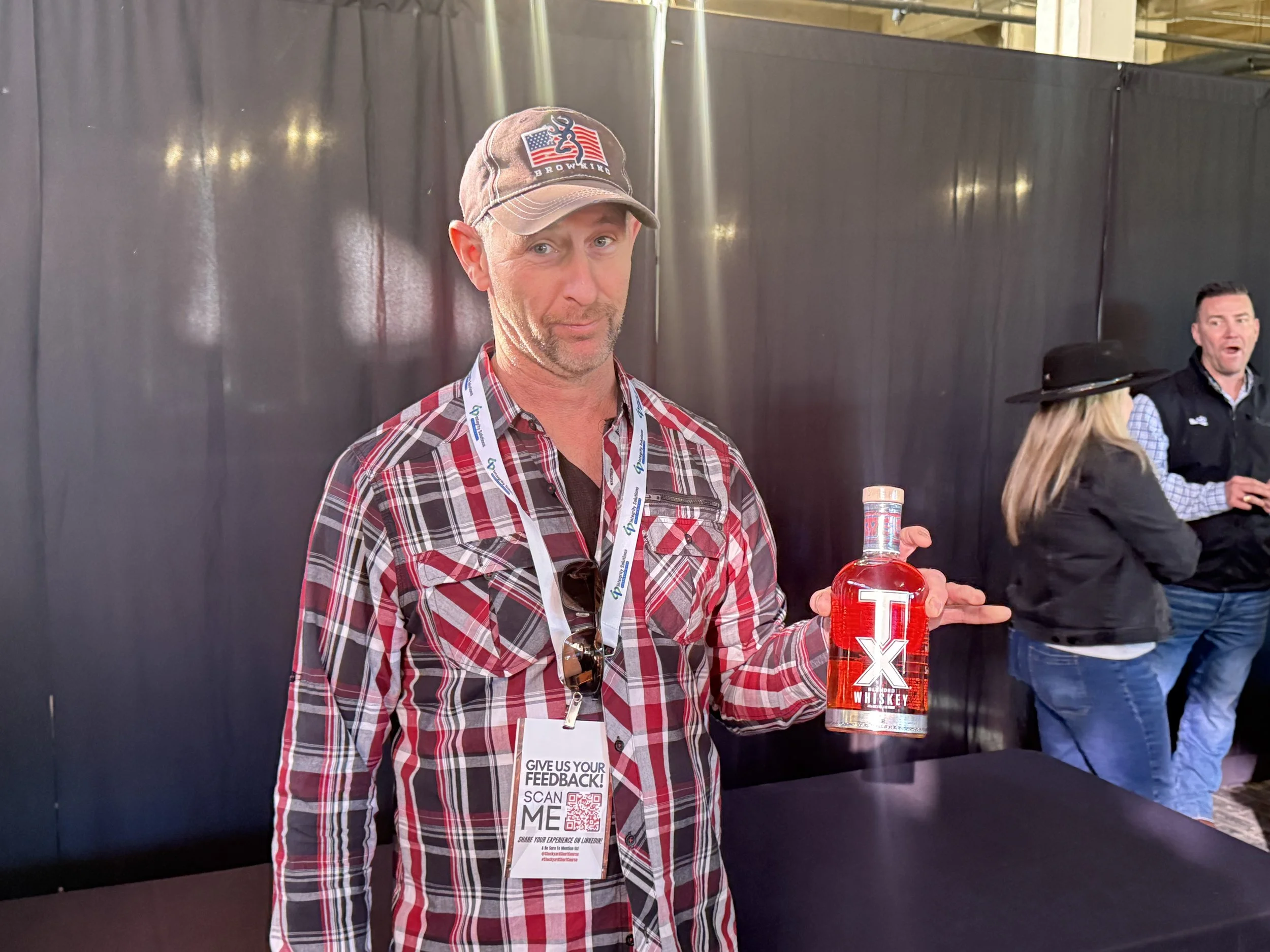 A man wearing a plaid shirt and a baseball cap holding a bottle of TX Whiskey at an indoor event, with two other people in the background.