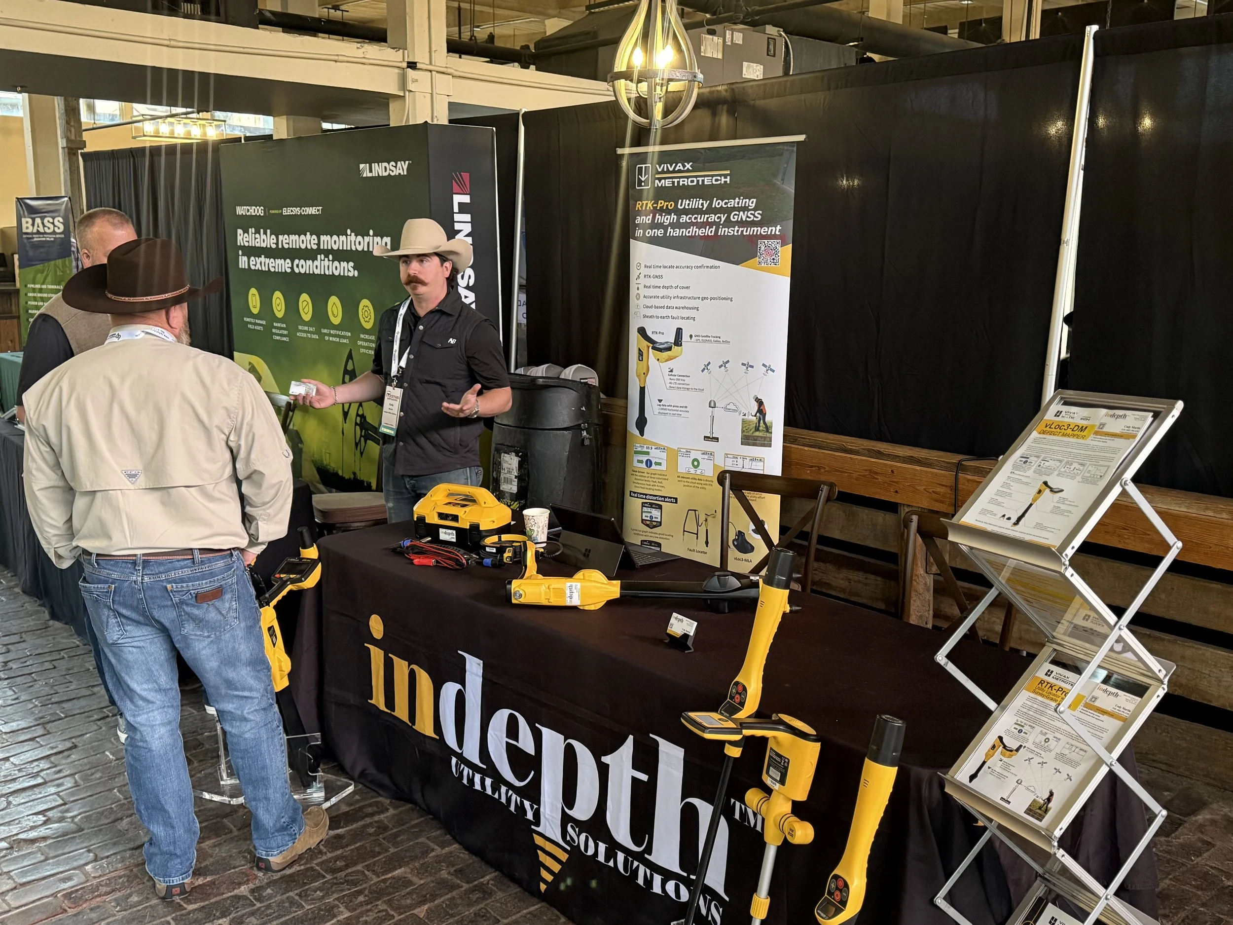 Trade show booth showcasing underground utility detection equipment, with a representative explaining features to two visitors. The booth displays tools, banners, and informational posters about GNSS and utility location technology.