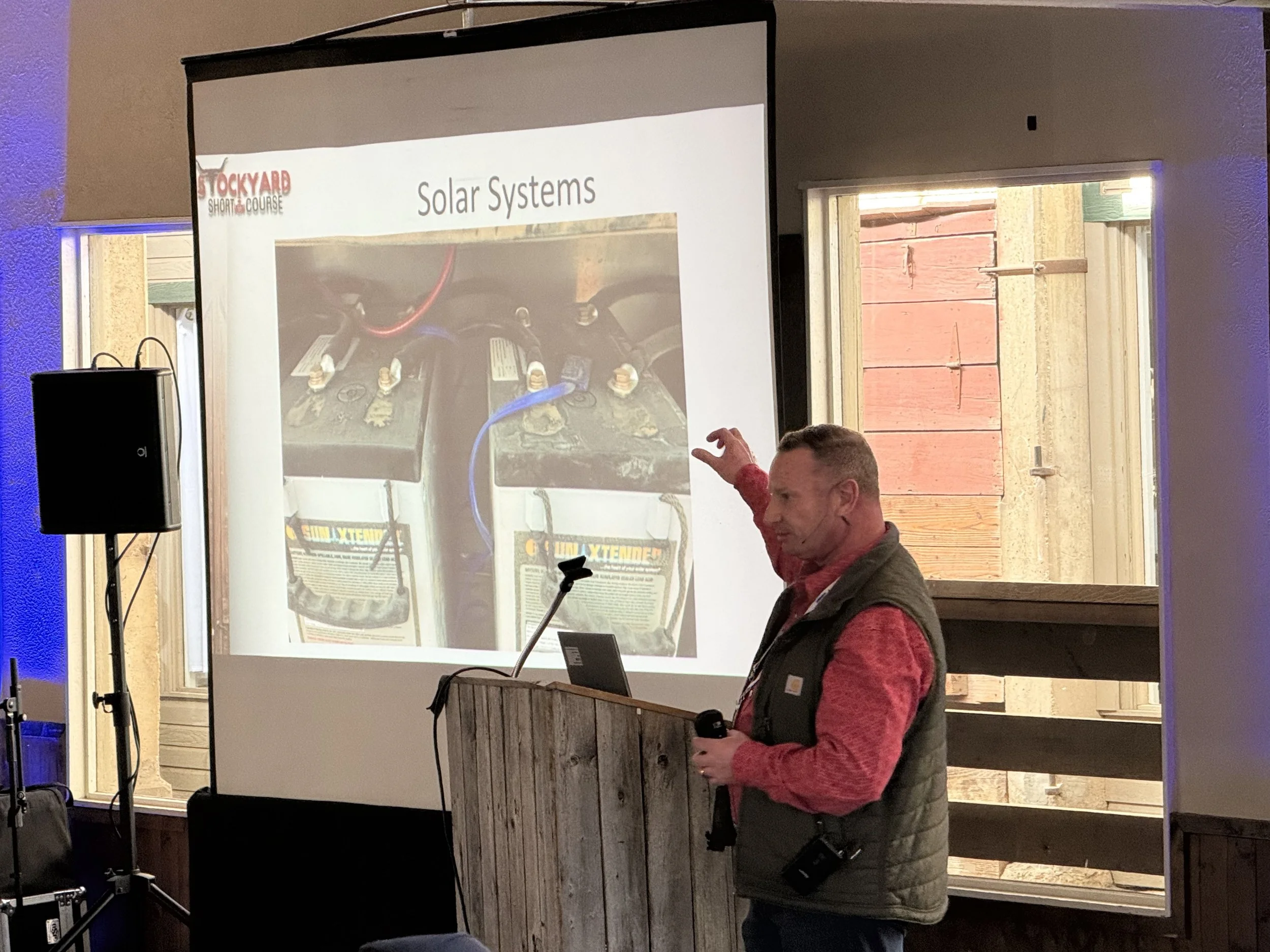 A man giving a presentation in front of a screen showing a slide about solar systems, with batteries and wires, at an indoor venue with wooden walls and a window.