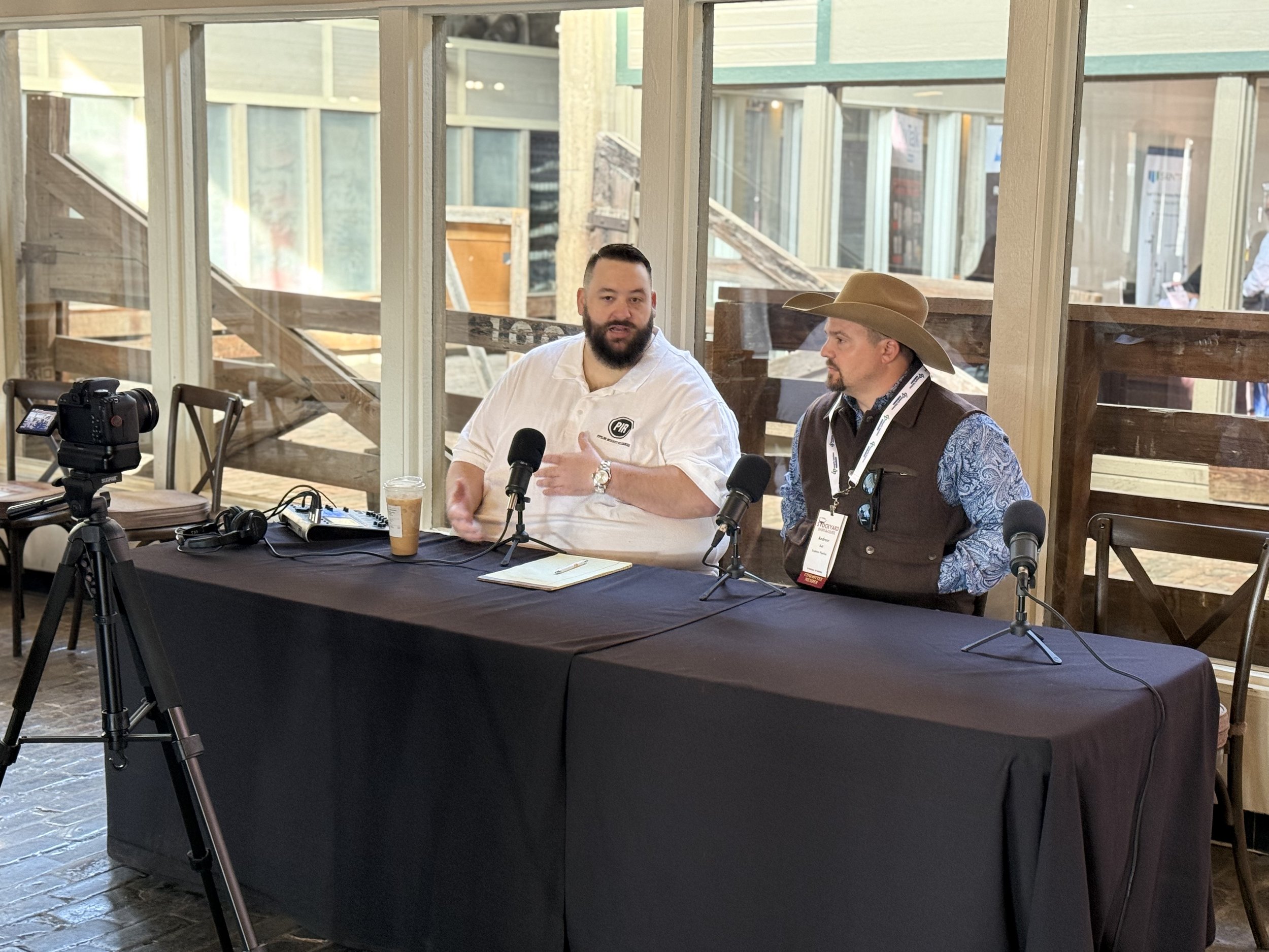Two men sitting at a black-covered table with microphones, a camera on a tripod, and a notepad, engaged in a discussion or interview, in a well-lit indoor space with glass windows and wooden fences outside.