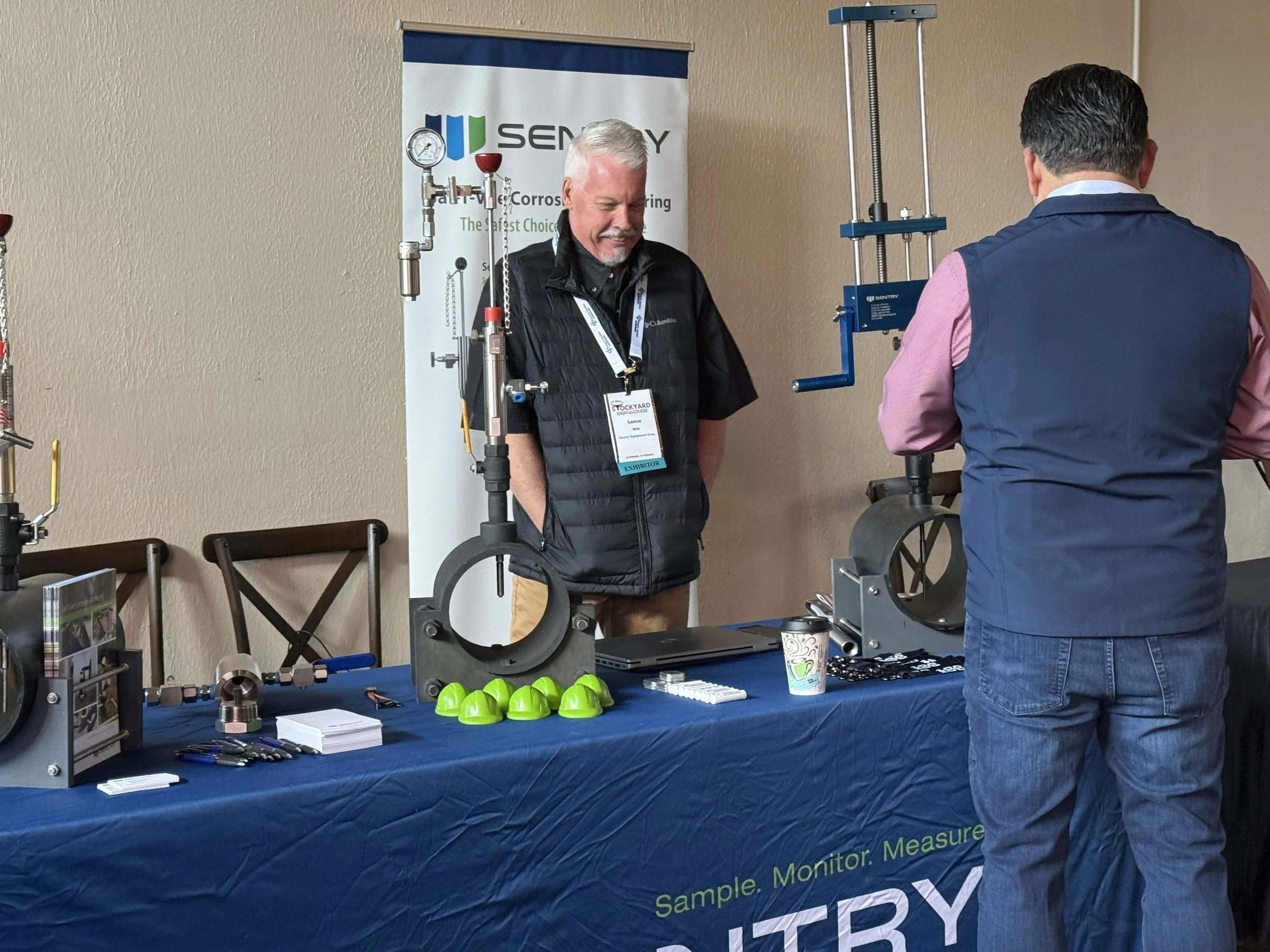 Two men at a booth with equipment and a Sentry Industries banner, engaging in a demonstration or discussion at a trade show or conference.