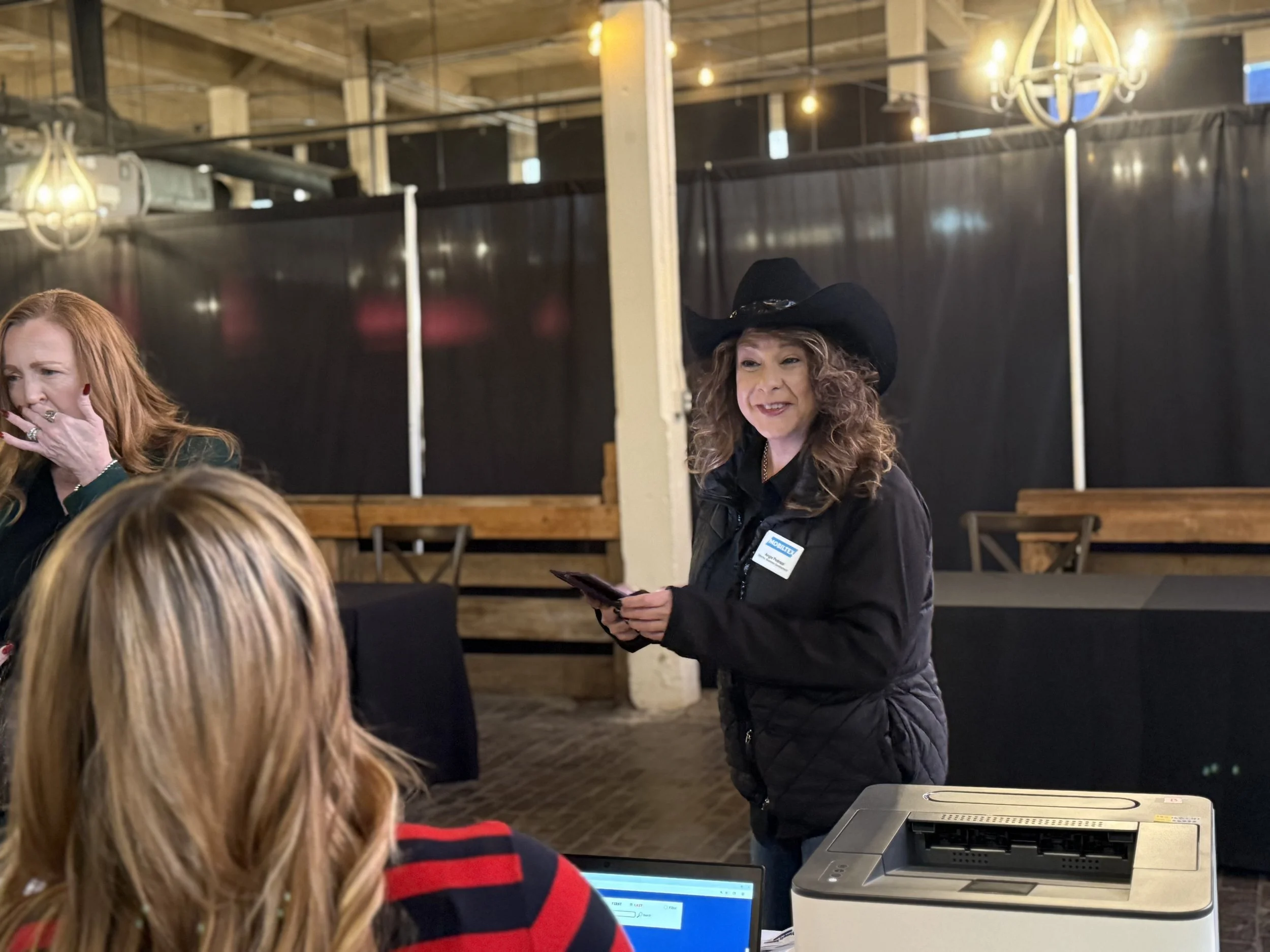 A woman wearing a black cowboy hat and jacket is holding a smartphone and talking to two women sitting at a table. One woman is wearing a red and black striped shirt, and the other has long red hair. There is a printer and a laptop on the table.
