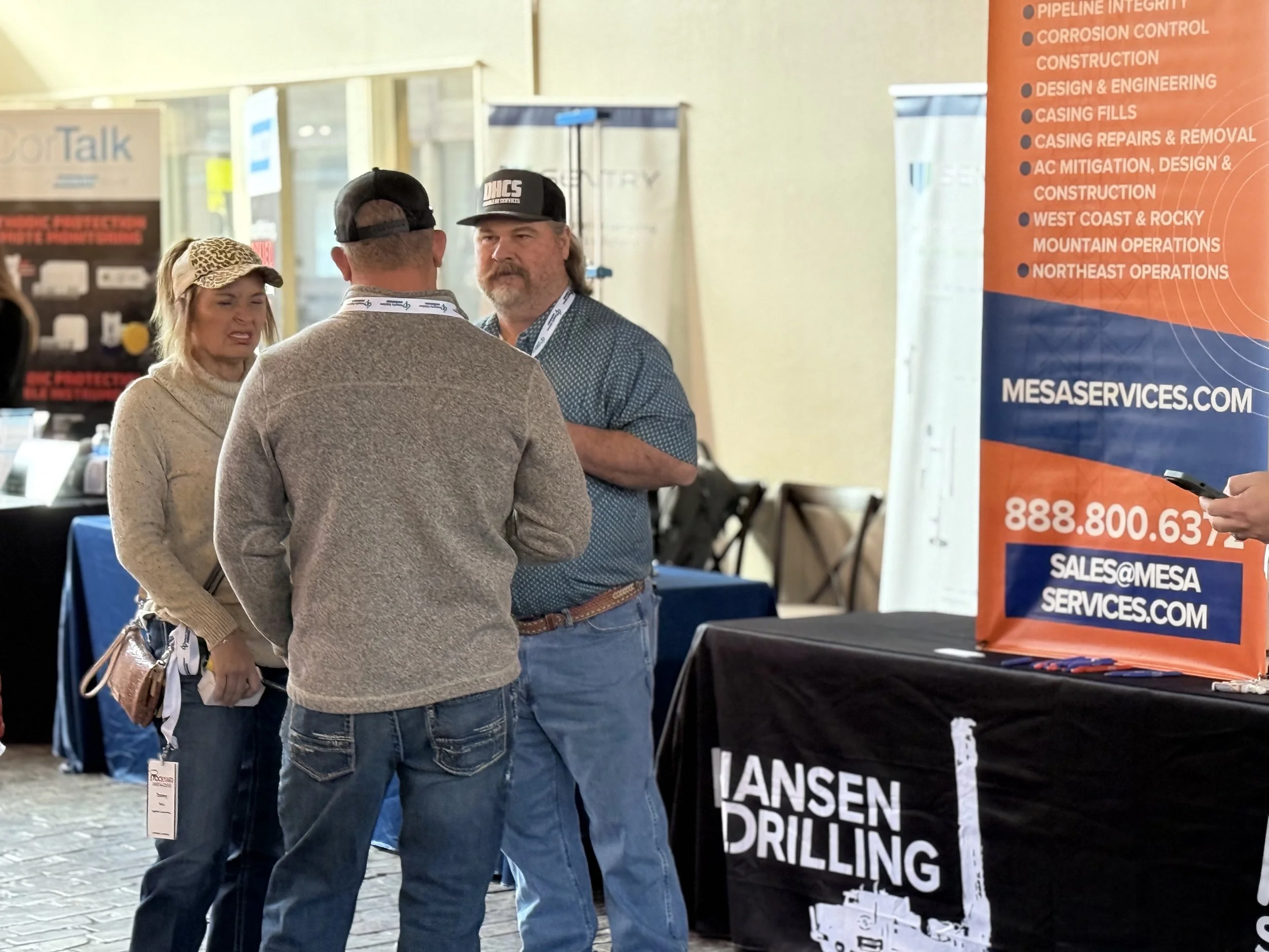 Three people are having a conversation at a trade show booth. The man facing away is wearing a gray sweater and cap, the woman is in a tan sweater and leopard print hat, and the man with a beard is in a blue shirt. There is a large orange banner to t