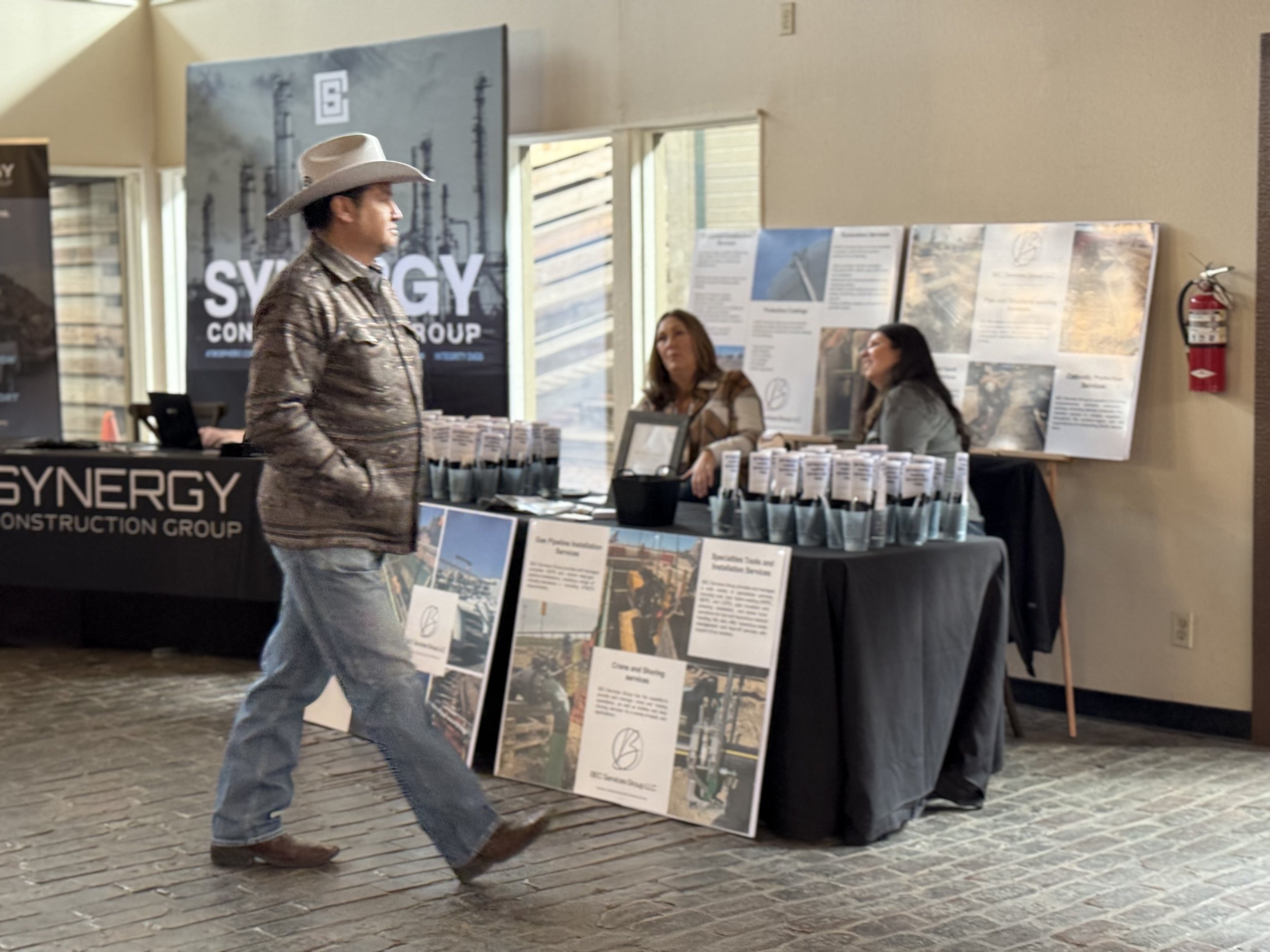 A man wearing a cowboy hat and western-style shirt walking past a booth for SYNERGY Construction Group at an indoor event.