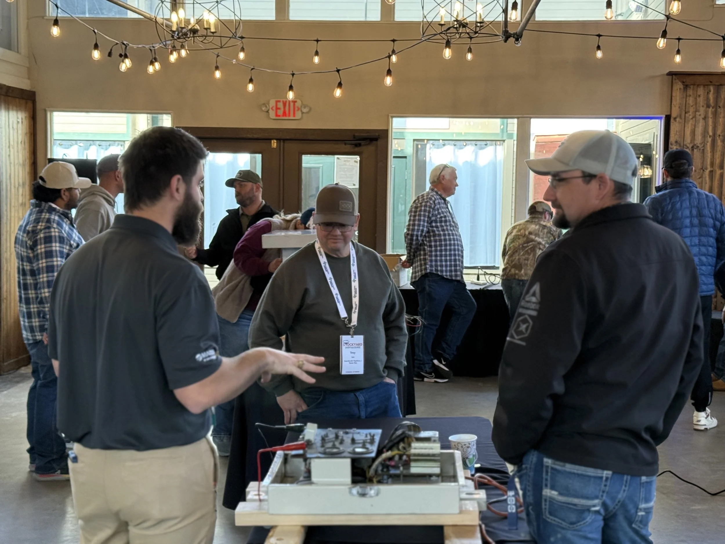 A group of men is gathered inside a building with string lights hanging from the ceiling. They are engaged in conversation around a table with electronic equipment on it. The background shows other people standing and talking near large windows, with