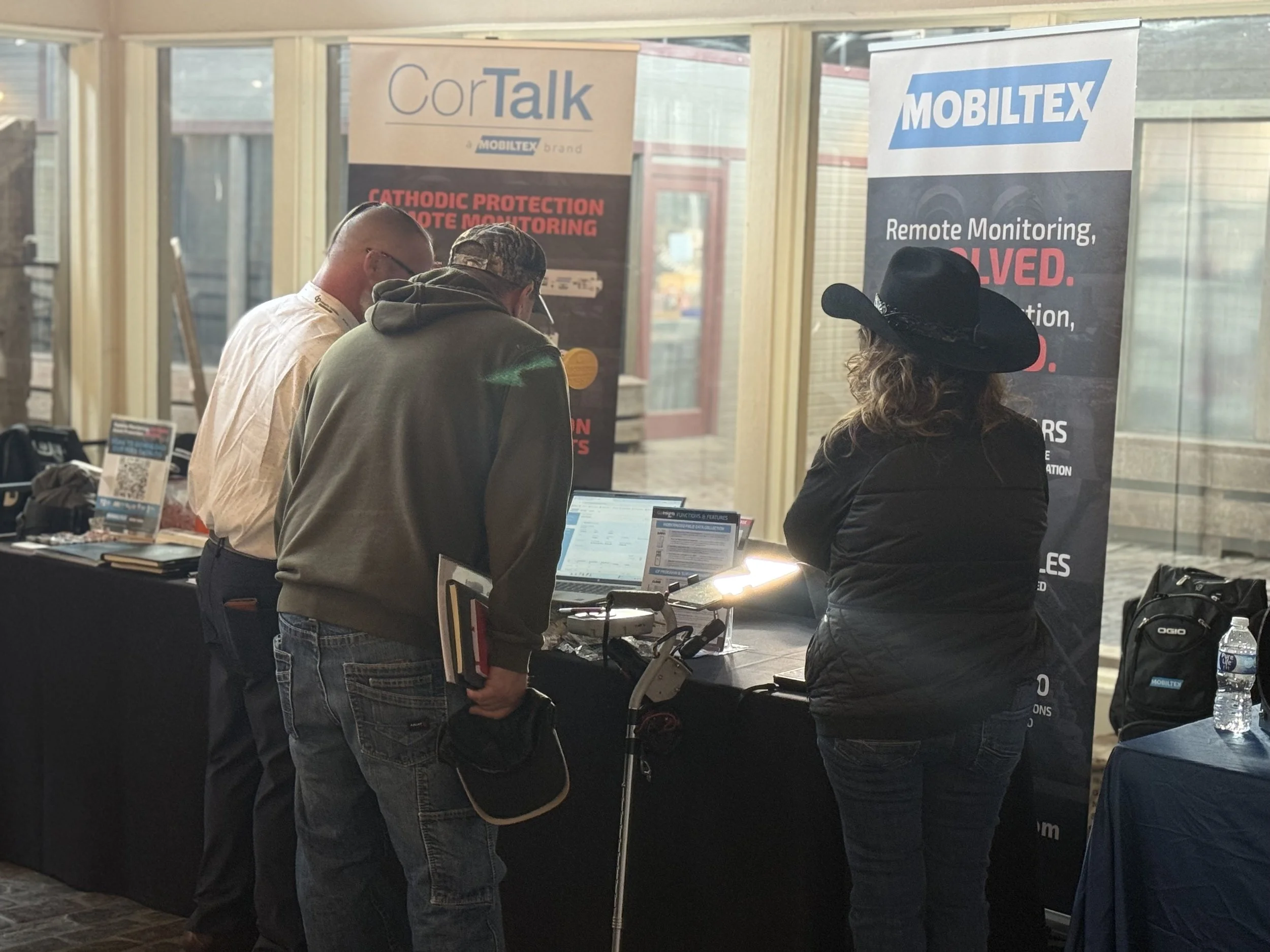 Three people at a booth looking at monitors, with banners displaying the company names 'CorTalk' and 'Mobiltex' and information about remote monitoring and active monitoring services.