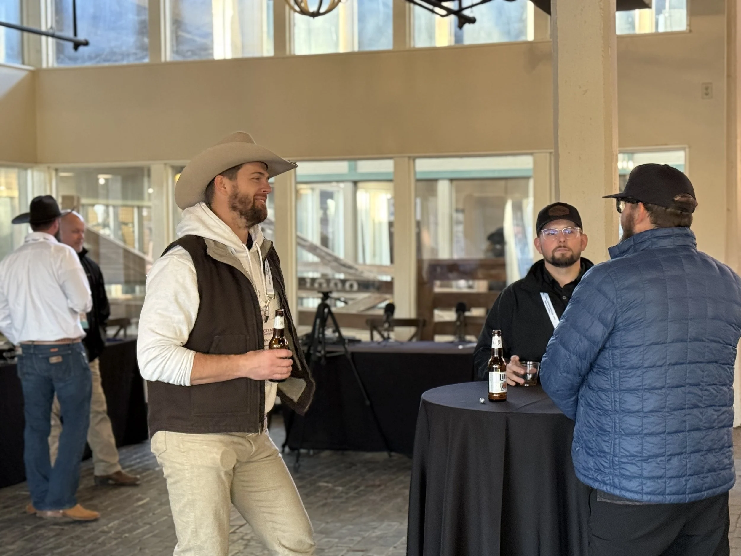 Three men are standing near a tall round table, holding beer bottles, and conversing indoors with large windows in the background. Two men are facing each other, and one man is standing to the side.