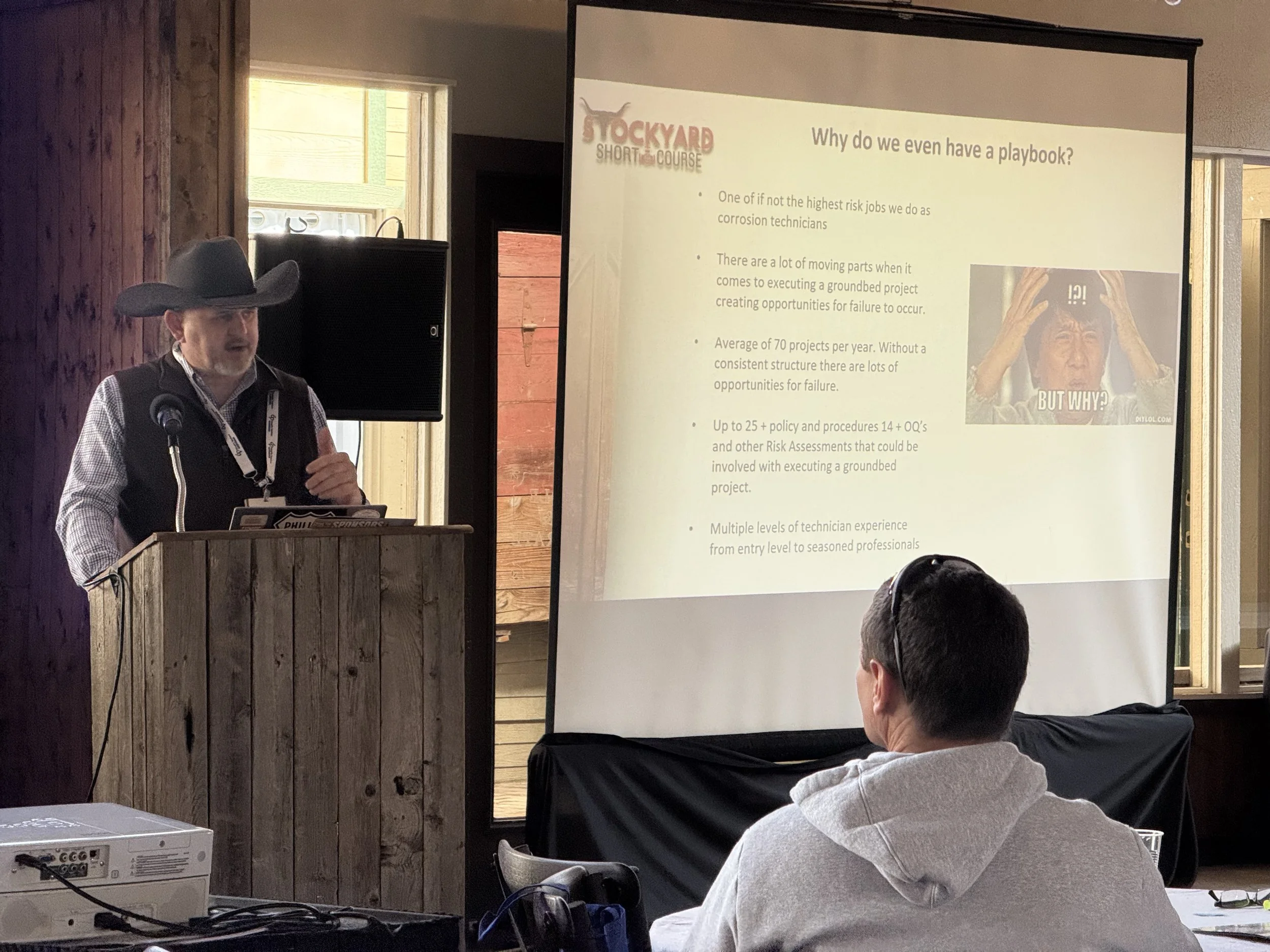 A man in a cowboy hat and vest giving a presentation at a conference. He is standing at a wooden podium with a large projection screen beside him. The slide on the screen discusses the reasons for having a playbook, with bullet points and an image of