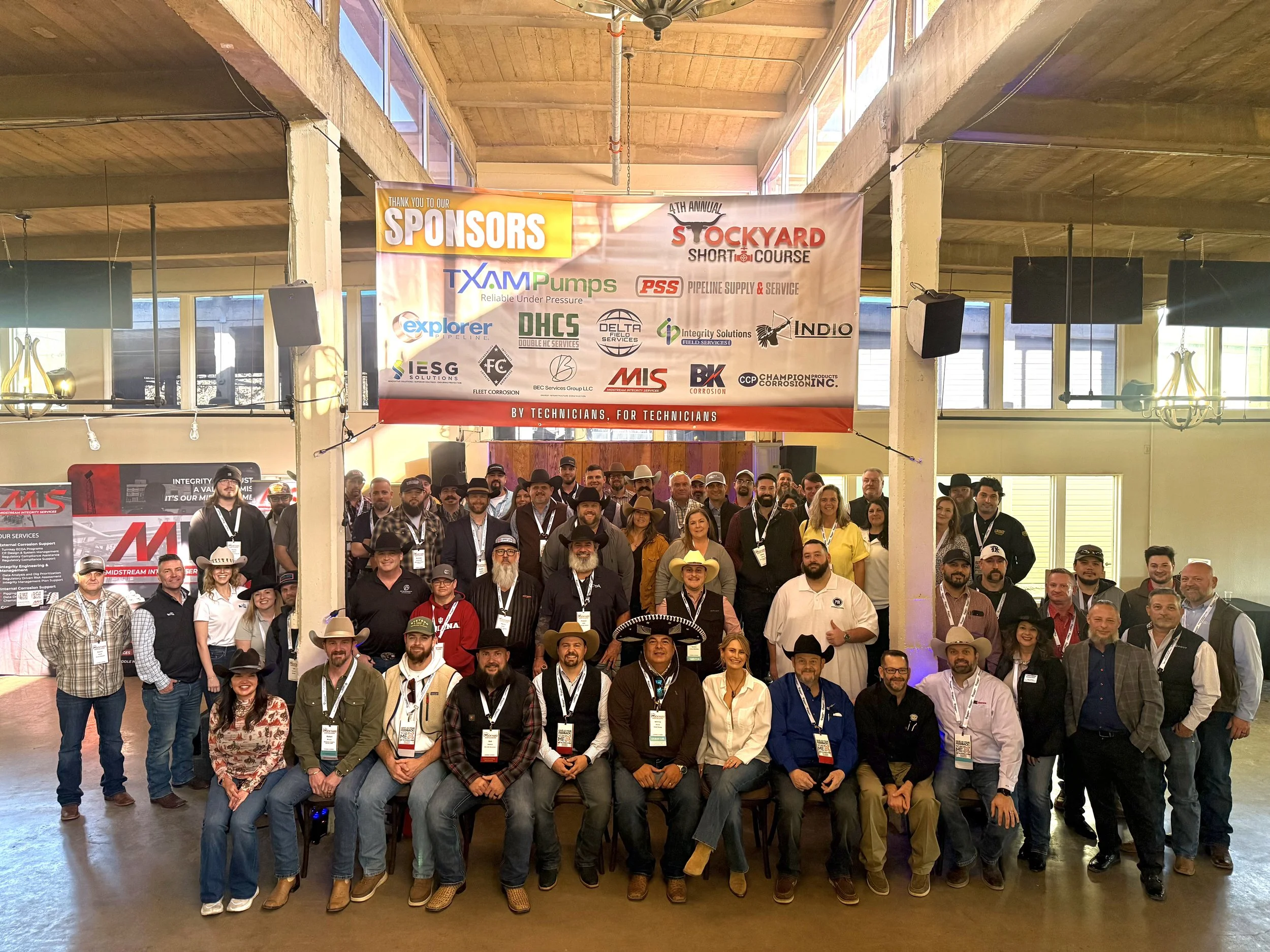 Group of people gathered indoors for a photo at a conference or event, with a large banner overhead displaying sponsor logos and event details.