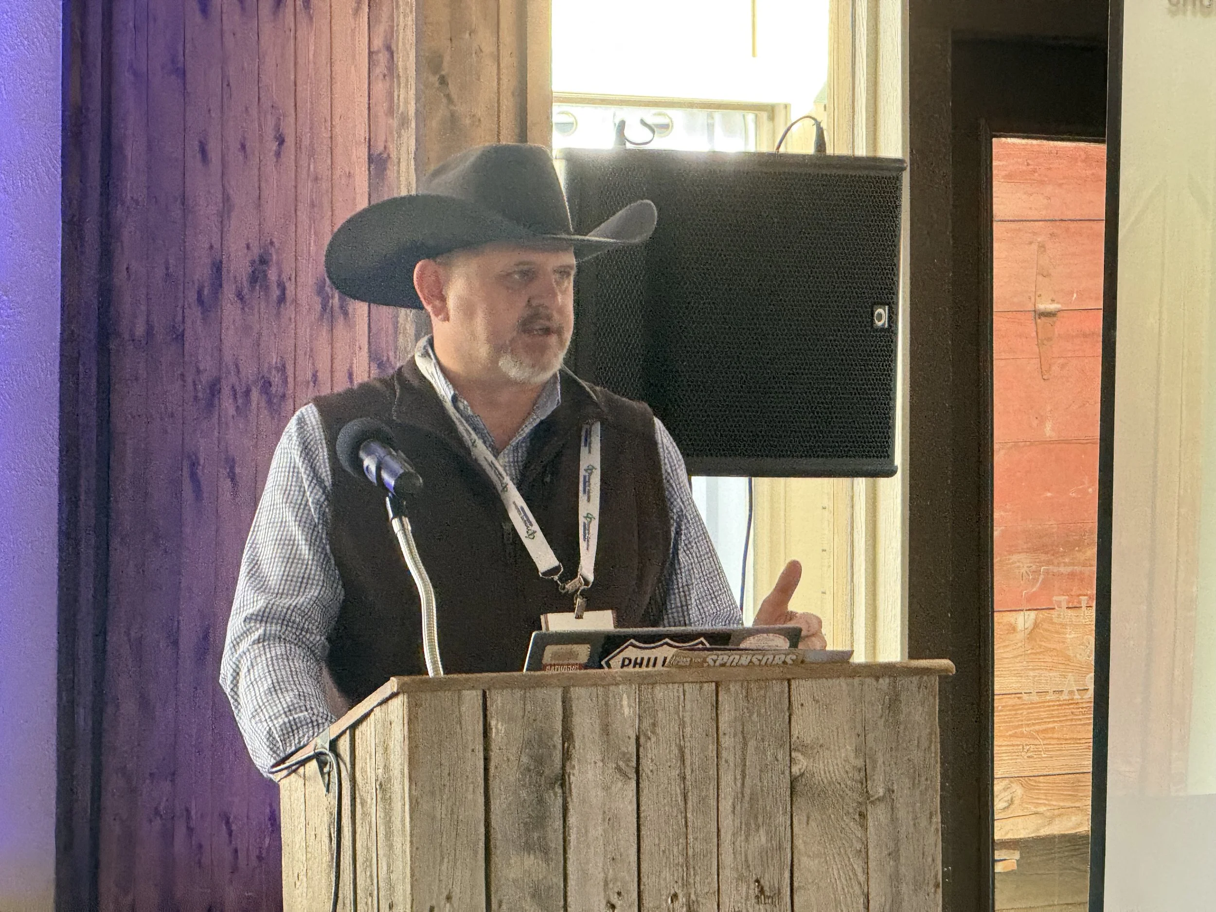 A man wearing a cowboy hat, plaid shirt, and dark vest standing behind a rustic wooden podium, speaking into a microphone, with a large speaker behind him in a wooden interior setting.