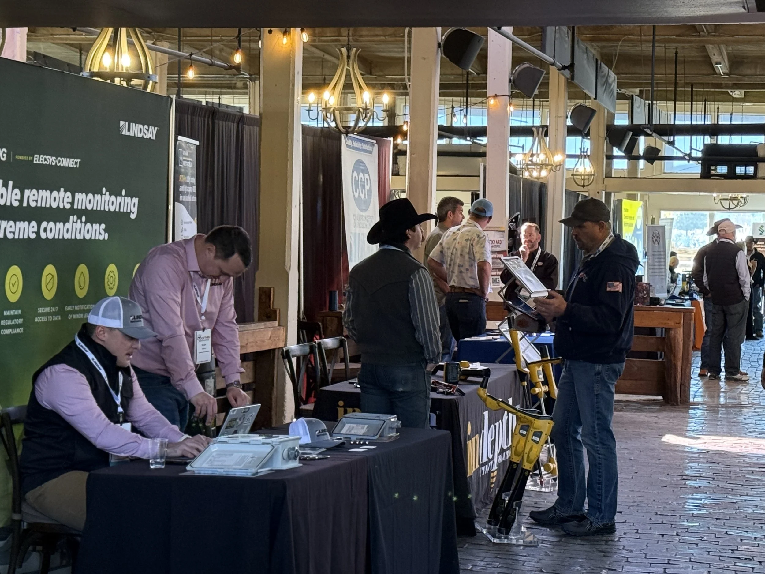At a trade show, several people are gathered around booths with displays and promotional materials. Some are talking to representatives, and there are various electronic devices and signs visible.