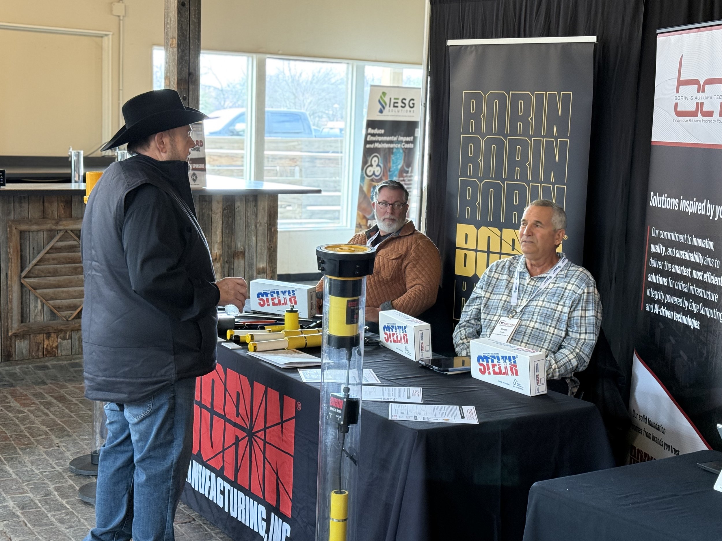 A man wearing a black cowboy hat and jacket is talking to two men seated at a booth at an event. The seated men are behind a table with various tools and equipment, and there are banners and posters in the background promoting different companies and