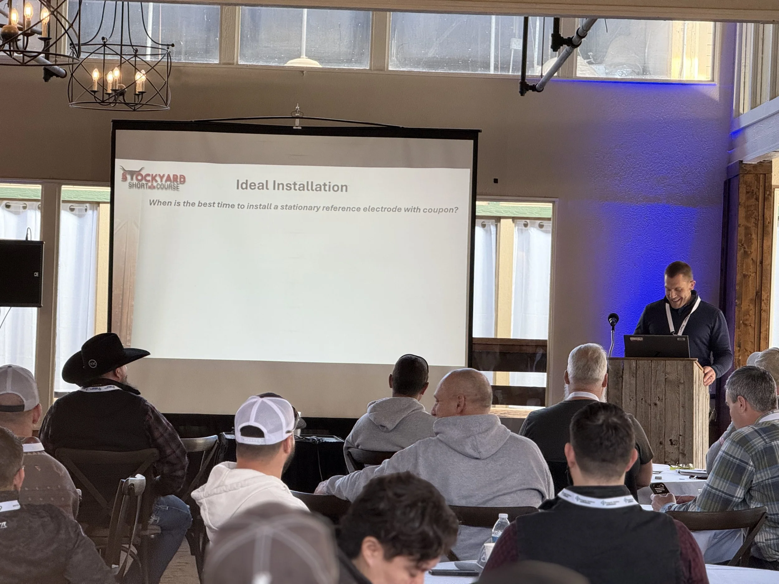 A man giving a presentation at a conference with a screen displaying a slide titled 'Ideal Installation' during a workshop. Attendees are seated, some taking notes, in a bright room with large windows.