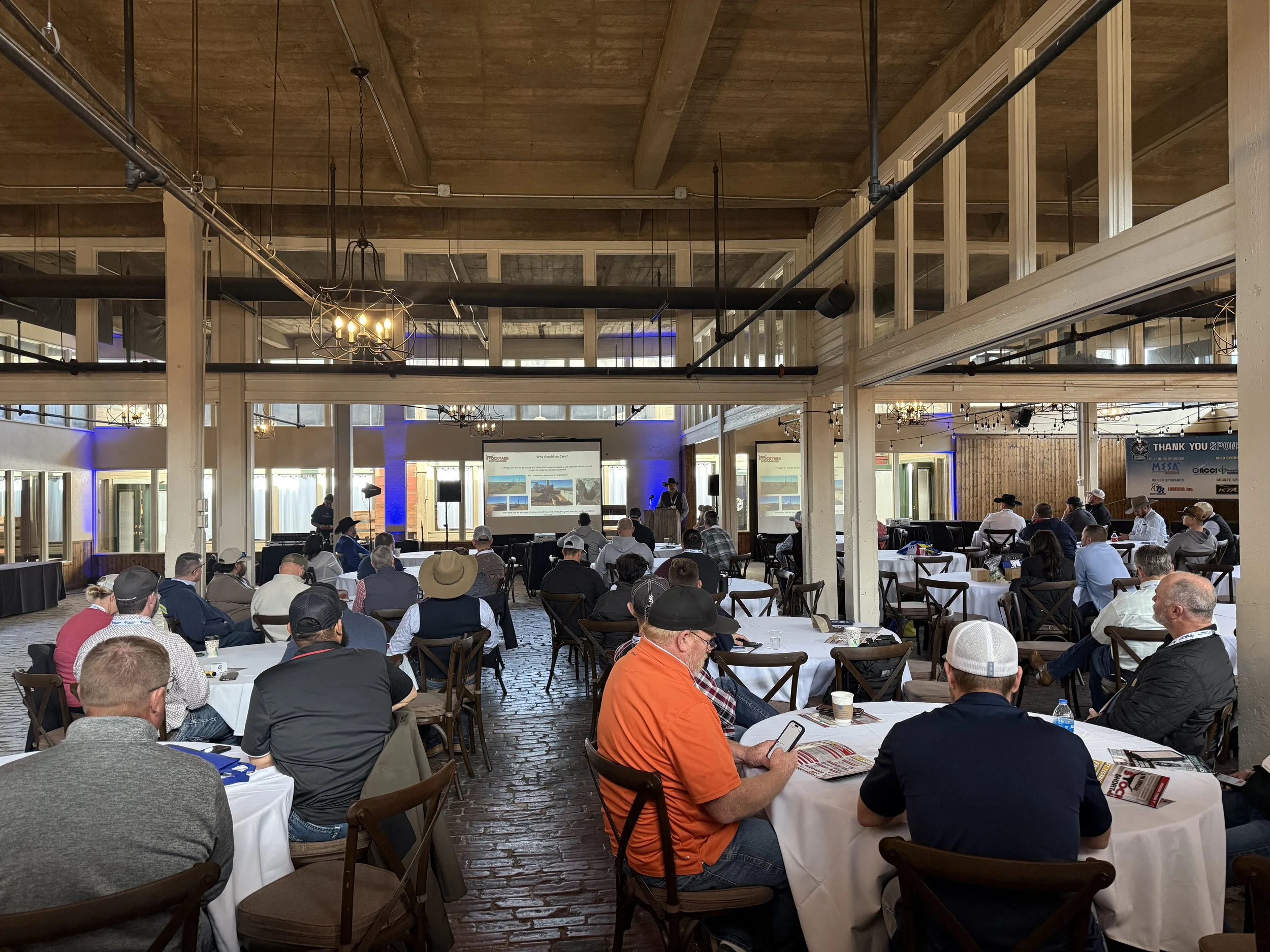 A conference or seminar with attendees seated at round tables listening to a speaker at a podium. The room has a rustic, industrial design with large windows, exposed beams, and chandeliers.