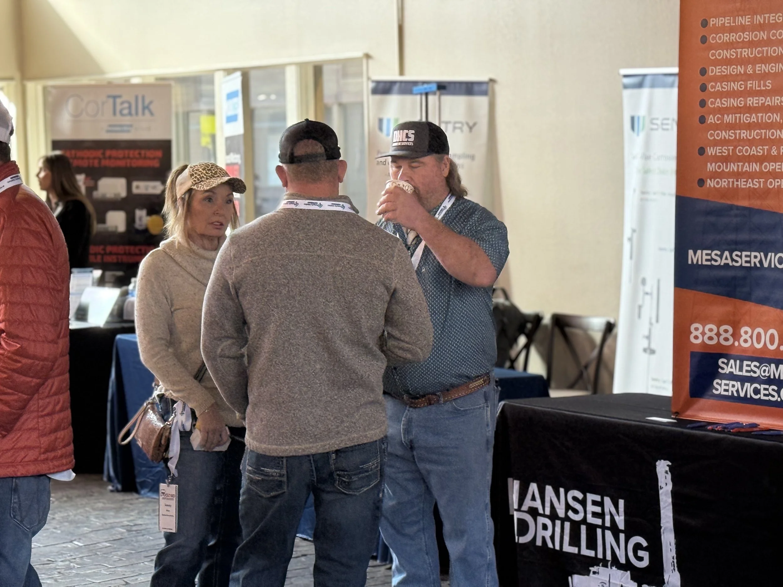 Three people talking at a trade show booth, one man drinking from a cup, with banners and displays in the background.