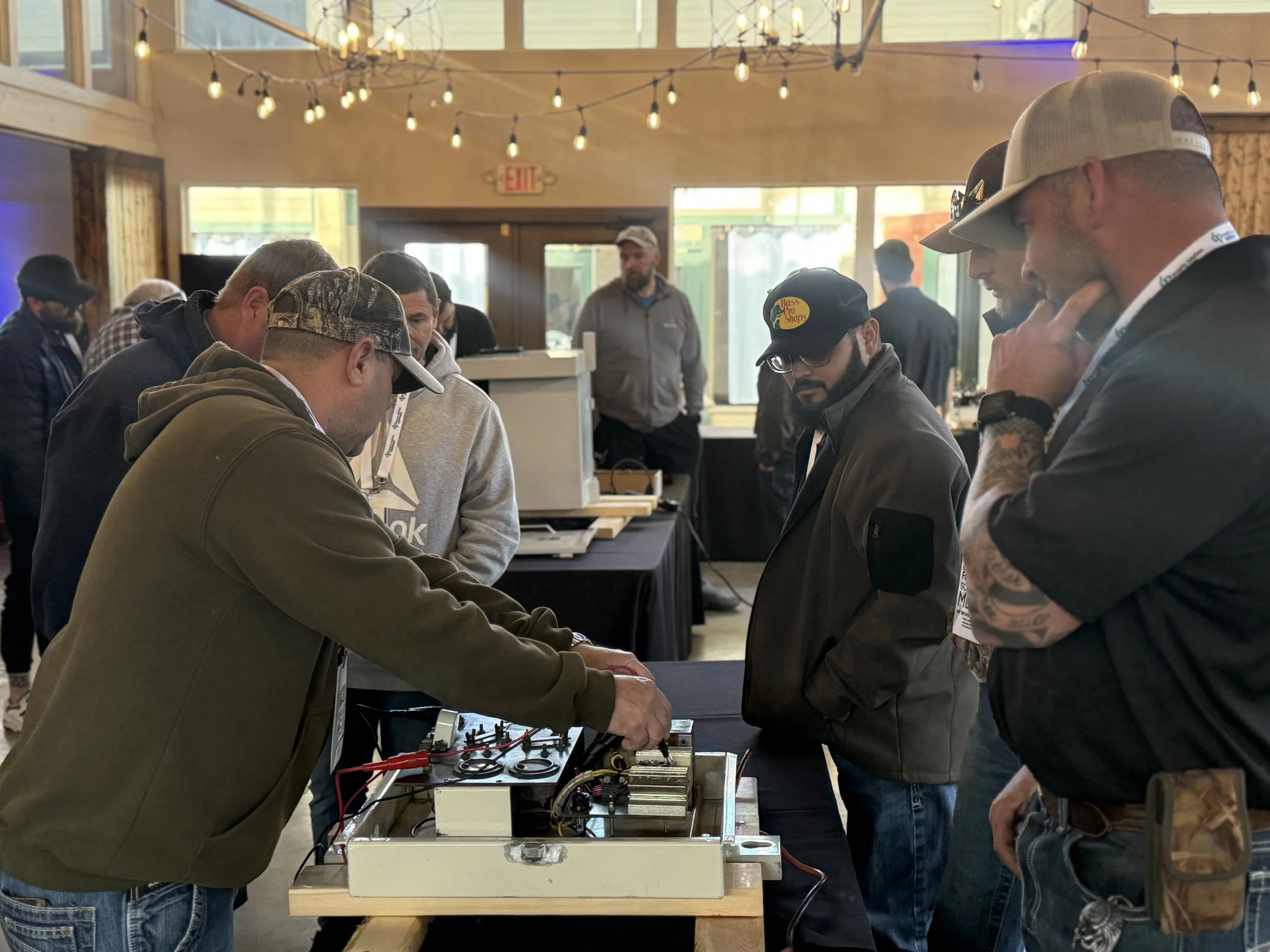 Group of men gathered around a workbench examining electronic equipment inside a well-lit room with string lights and large windows.
