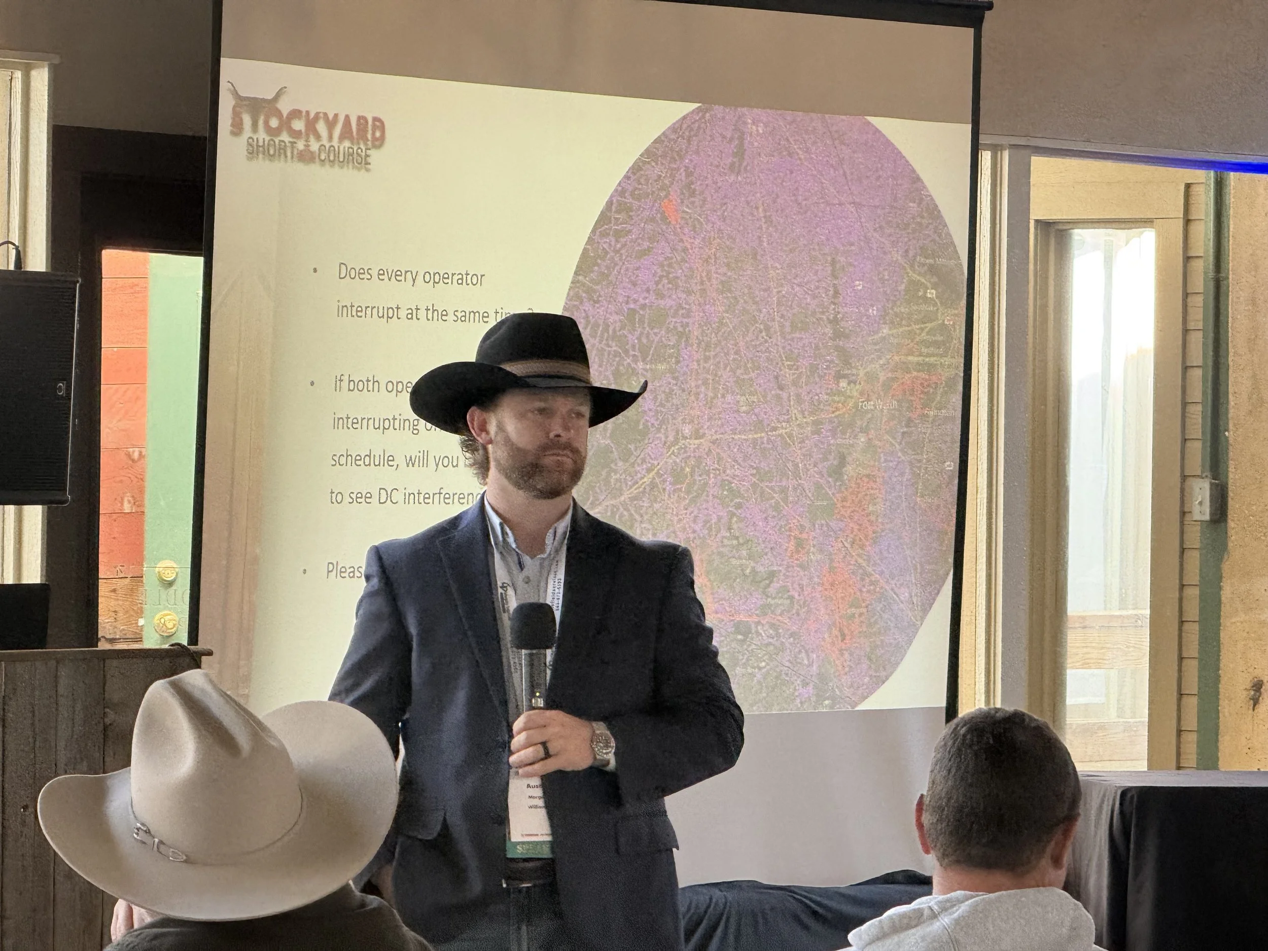 A man in a dark suit and white shirt, wearing a cowboy hat, standing in front of a screen with a presentation slide, holding a microphone. There is an audience with at least three visible members, one wearing a cowboy hat. The slide on the screen sho