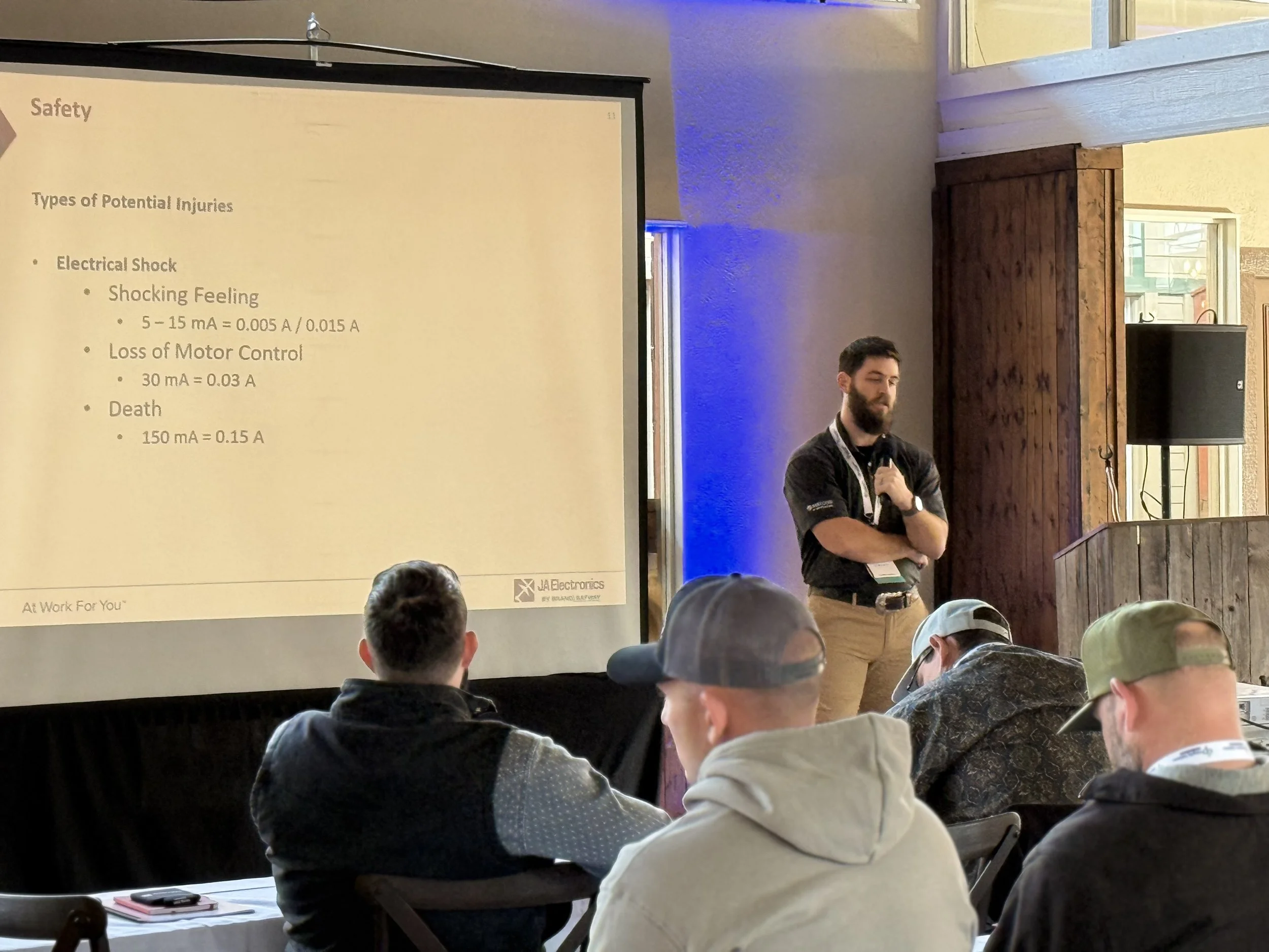 A man standing at a speaker's podium during a presentation with a large screen displaying safety information about electrical shocks and injuries in the background. The audience is seated in front, watching the presenter.