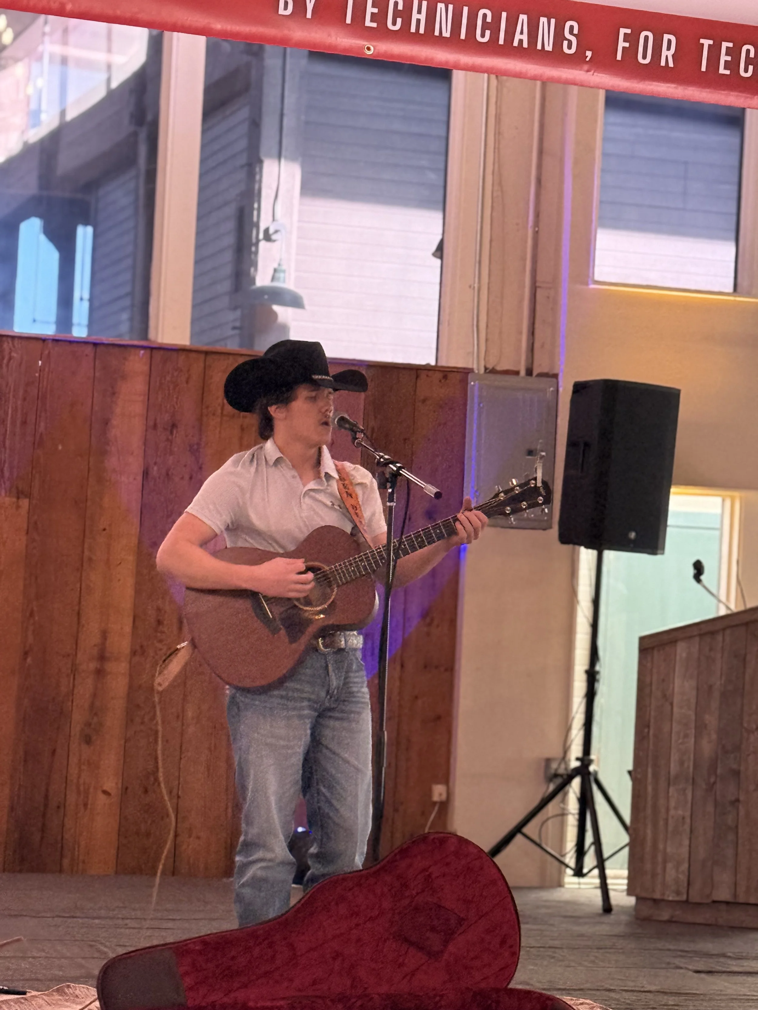 A man wearing a black cowboy hat, light-colored shirt, and jeans plays an acoustic guitar and sings into a microphone on a stage with wooden walls and a large speaker.