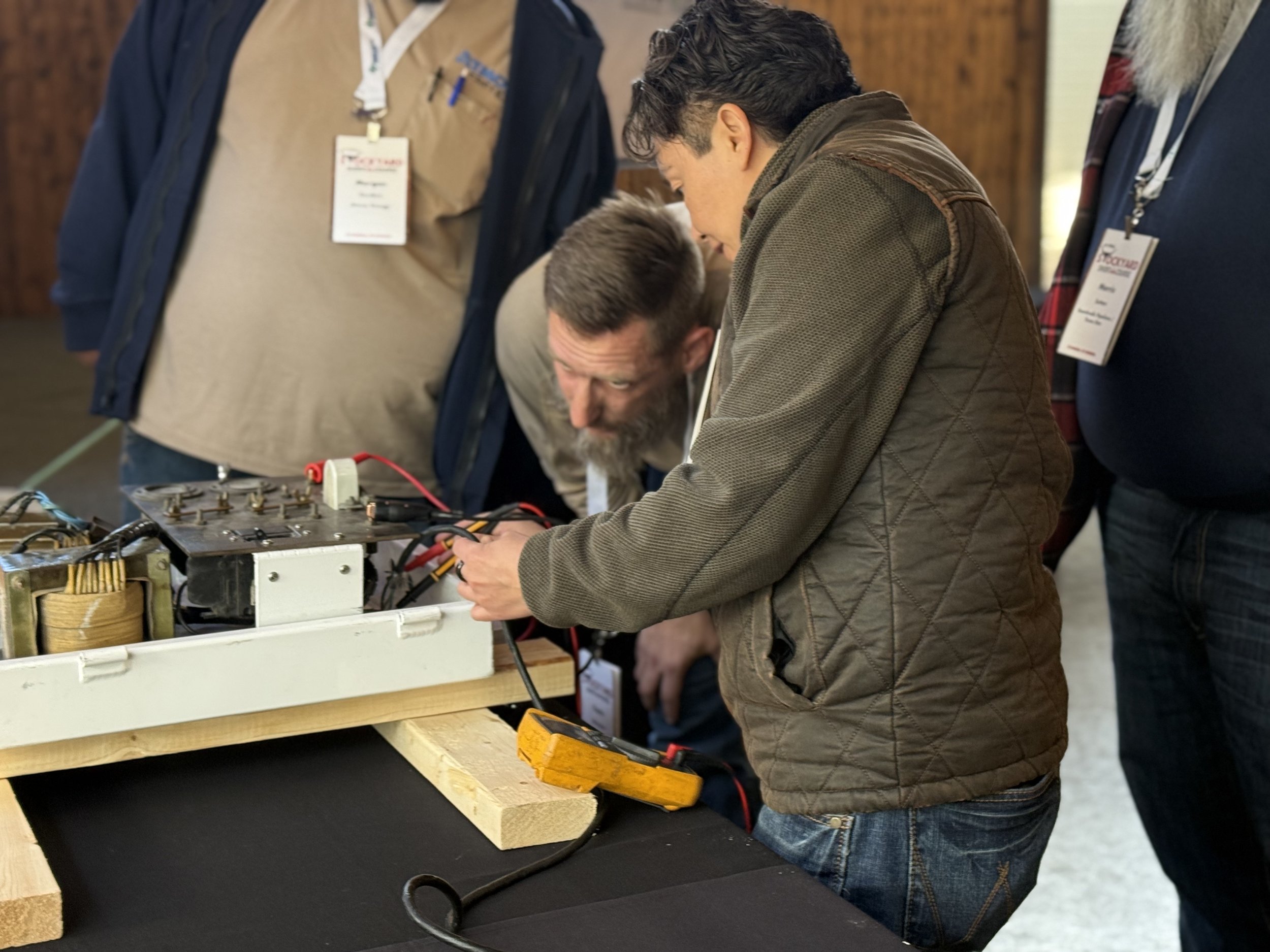 People working on electrical components at a workshop or exhibition. One person is using a multimeter on a setup with wires and equipment.