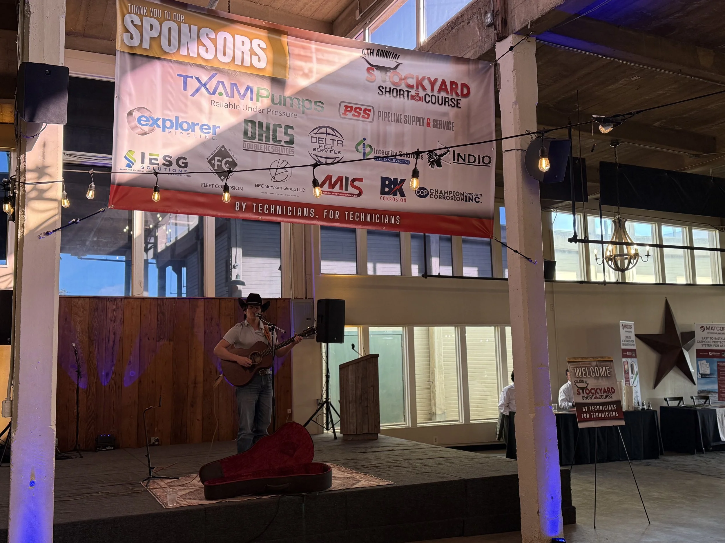 A person wearing a hat and white shirt playing guitar on a small stage, with a red open guitar case on the floor. Behind them is a large banner with sponsor logos, and to the right side, there are tables and a welcome sign for an event called Stockya