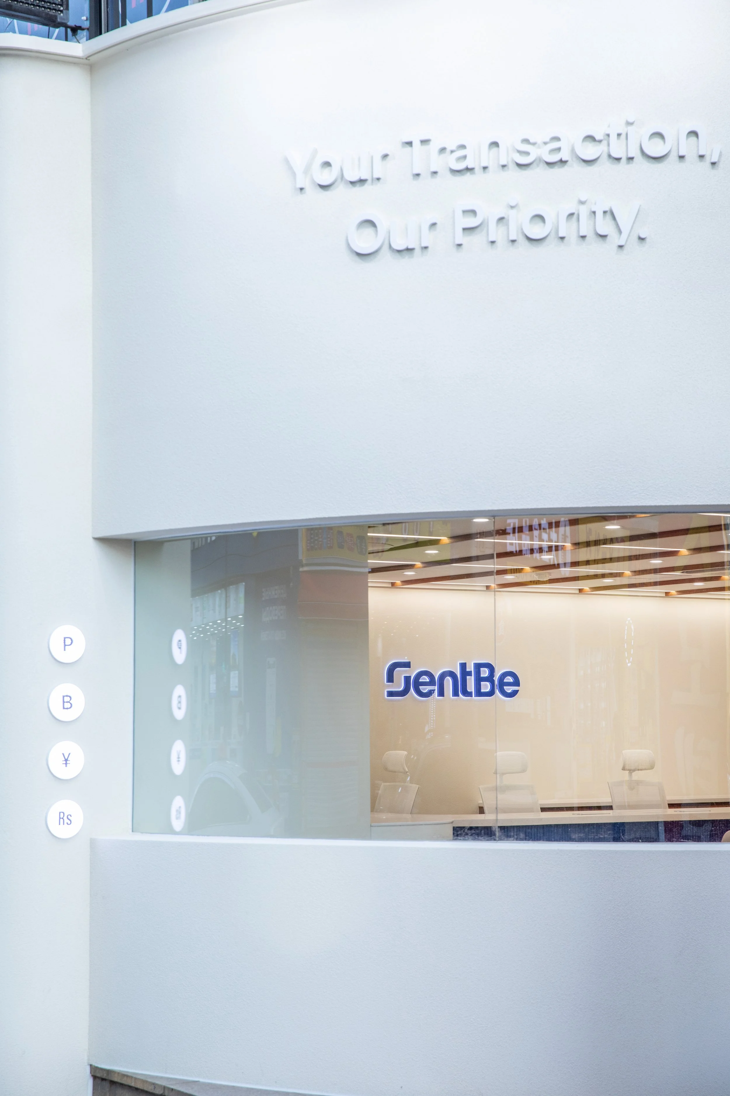 A modern banking or financial service branch with branding for 'SentBe'; the phrase 'Your Transaction, Our Priority' is displayed on the wall.