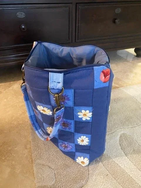 A blue tote bag with floral and lips patchwork design, standing on a beige carpeted floor in front of a dark wooden cabinet.