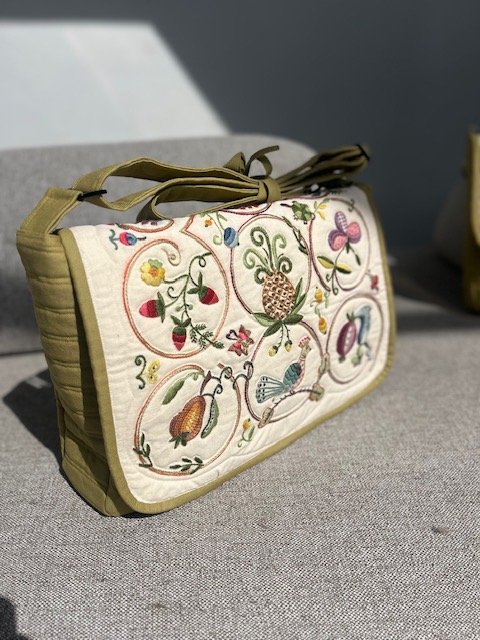 Beige embroidered fabric clutch with floral and butterfly designs, tied with a beige ribbon, resting on a gray surface.