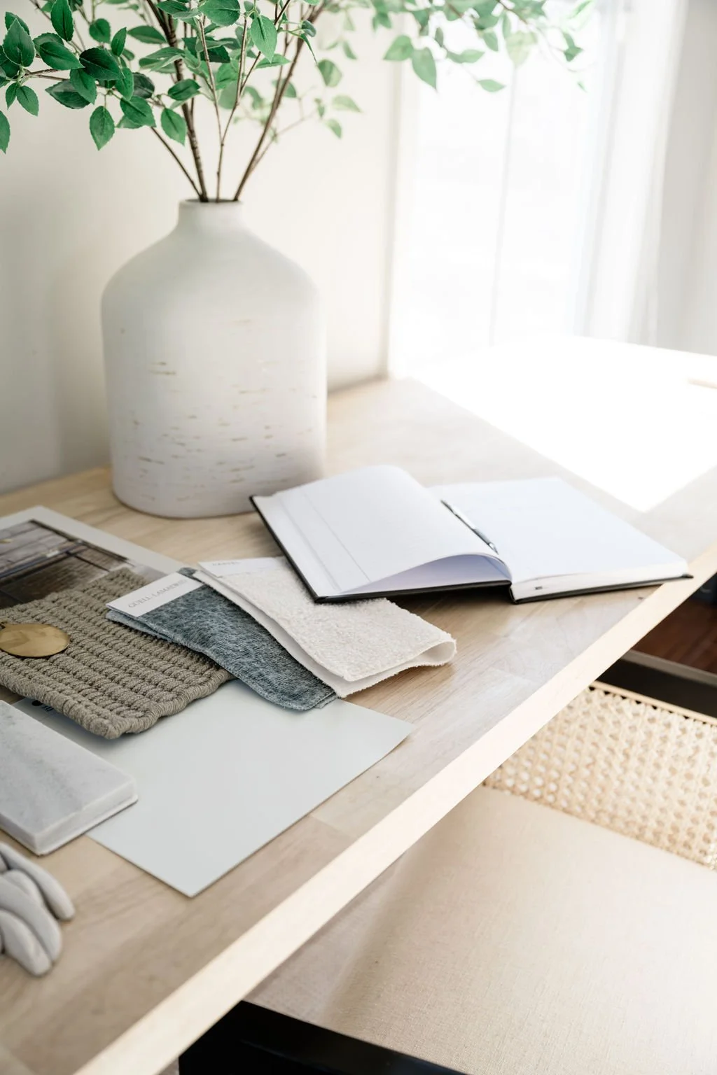 A wooden desk with fabric samples, a sketchbook, and a large white vase with green leafy branches near a window with sunlight.
