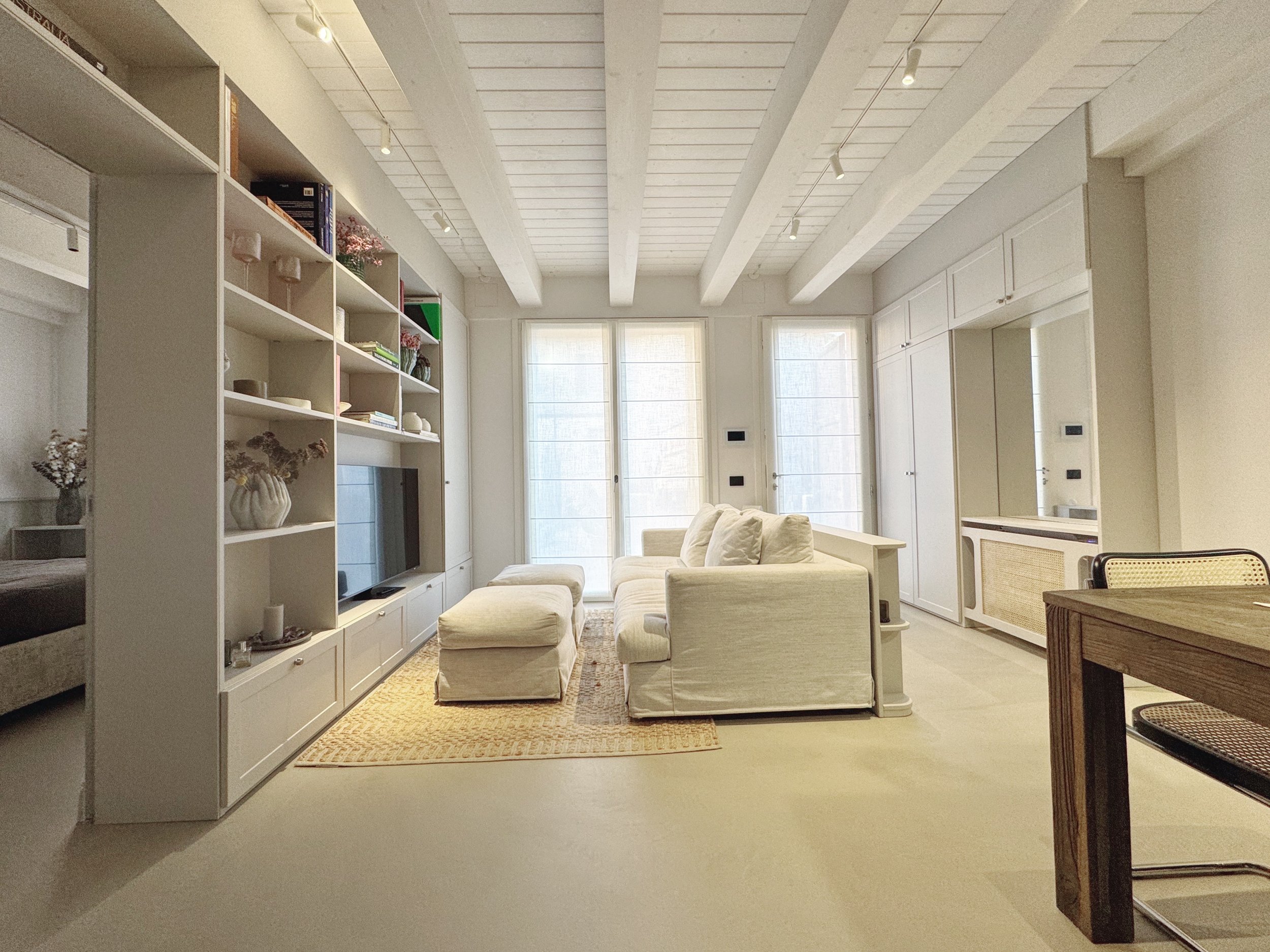 Salotto luminoso con divano bianco, televisore e scaffale con decorazioni e libri, arredamento minimalista con pareti bianche, finestre grandi con tende trasparenti.