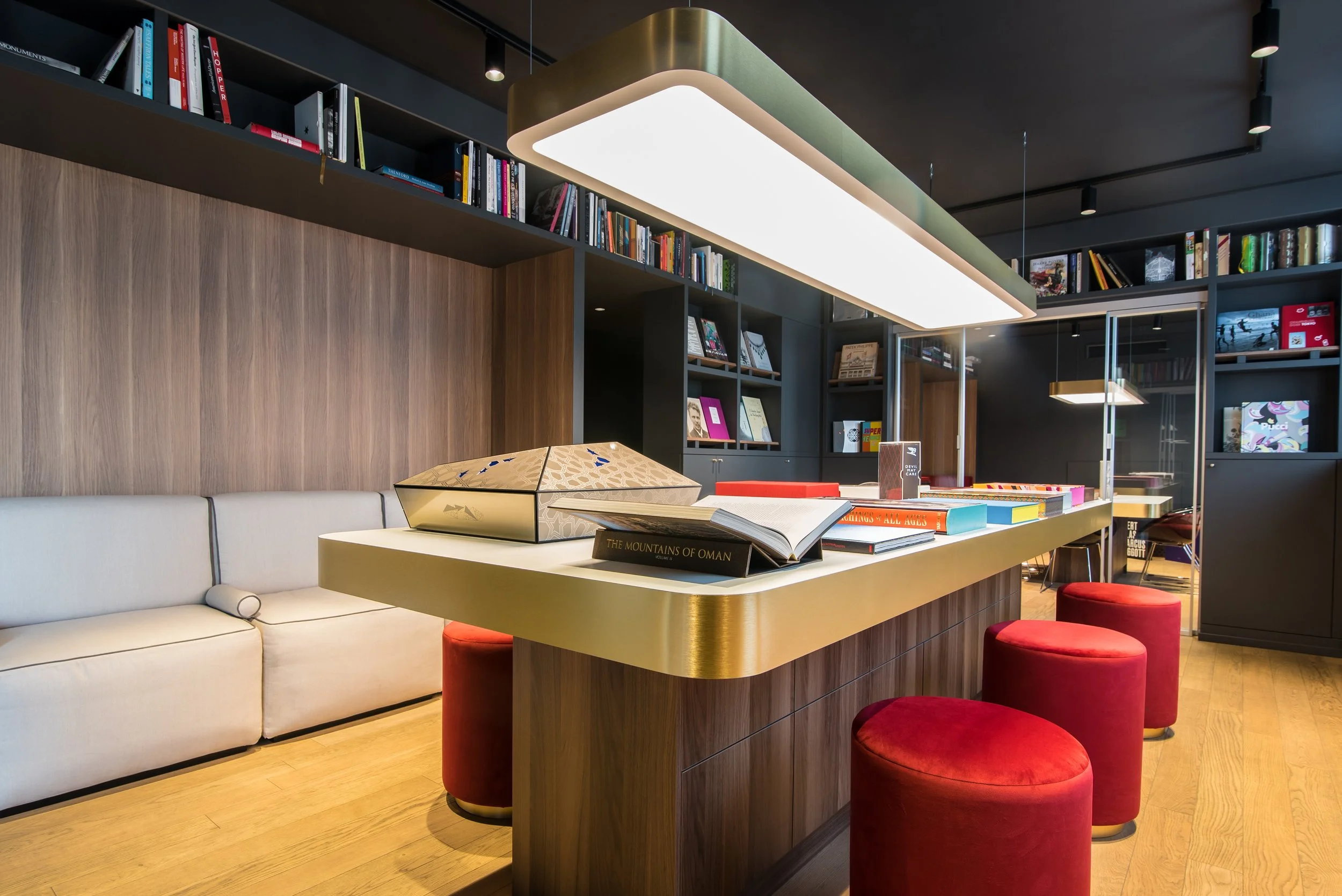 Interno di una libreria con uno scaffale pieno di libri, un tavolo con libri aperti e vari oggetti, sedie rosse e un divano bianco, illuminato da luci moderne.