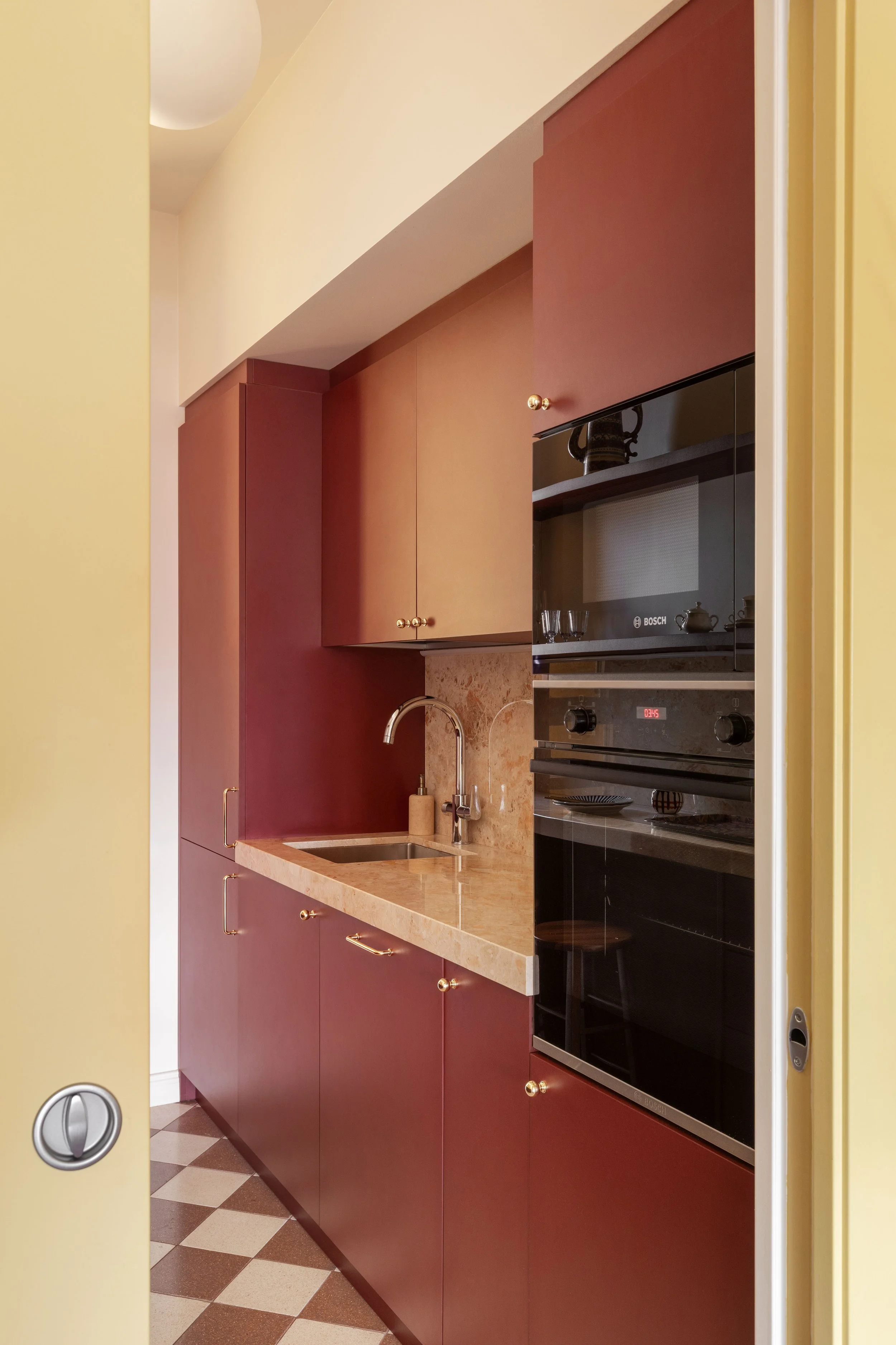 Cucina con armadietti marroni, forno elettrico Bosch, lavabo con getto d'acqua, pareti gialle e pavimento a scacchi beige e marrone.