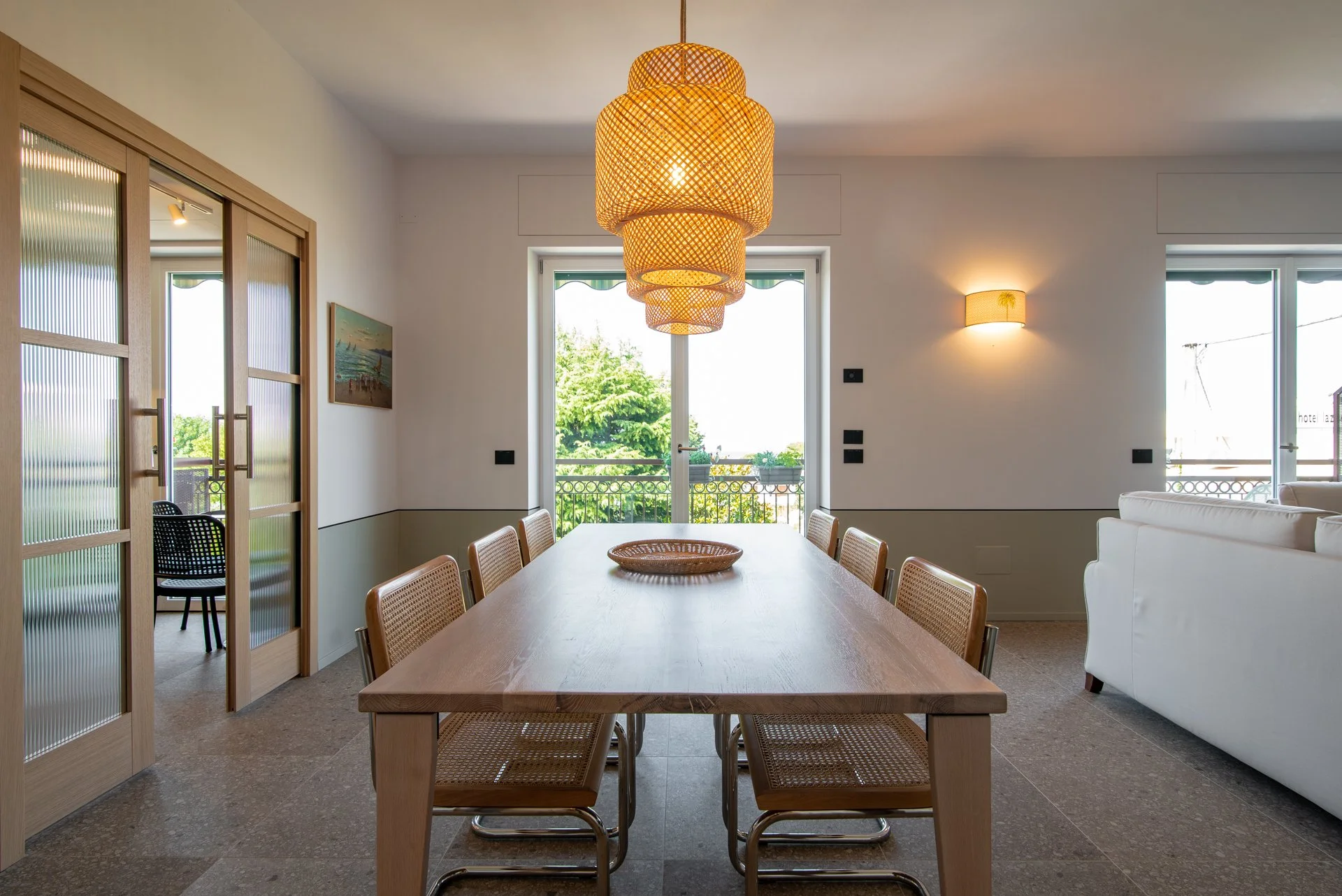 Interno di una sala da pranzo con tavolo in legno, sedie in legno e fibra naturale, illuminazione sospesa di design, porte a vetri e balcone con vista verde.