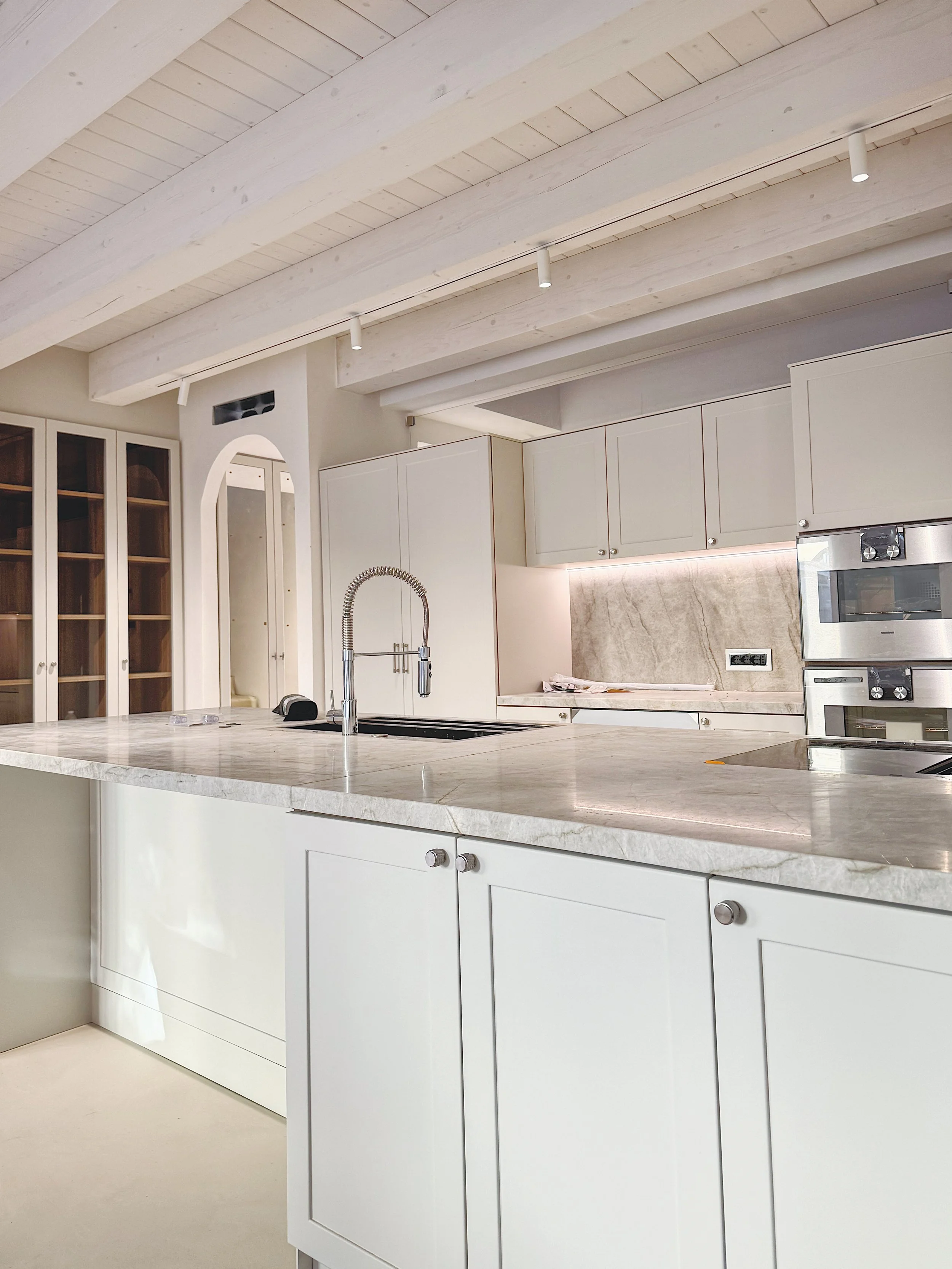 Cucina moderna con superfici in marmo chiaro, armadietti bianchi e soffitto con grandi travi in legno dipinte di bianco. C'è un lavandino con un rubinetto moderno e un forno integrato.