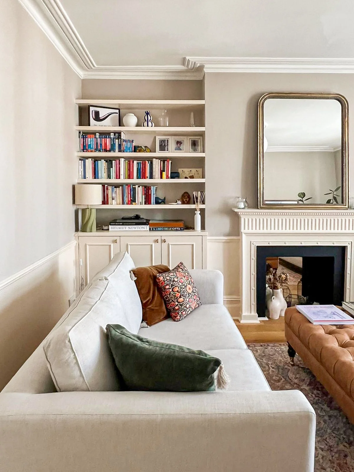 Soggiorno con camino bianco, grande specchio sopra il camino, scaffale con libri, divano beige con cuscini verdi, marroni e uno con motivo floreale, tavolino con riviste, pareti color crema e soffitto con cornice.