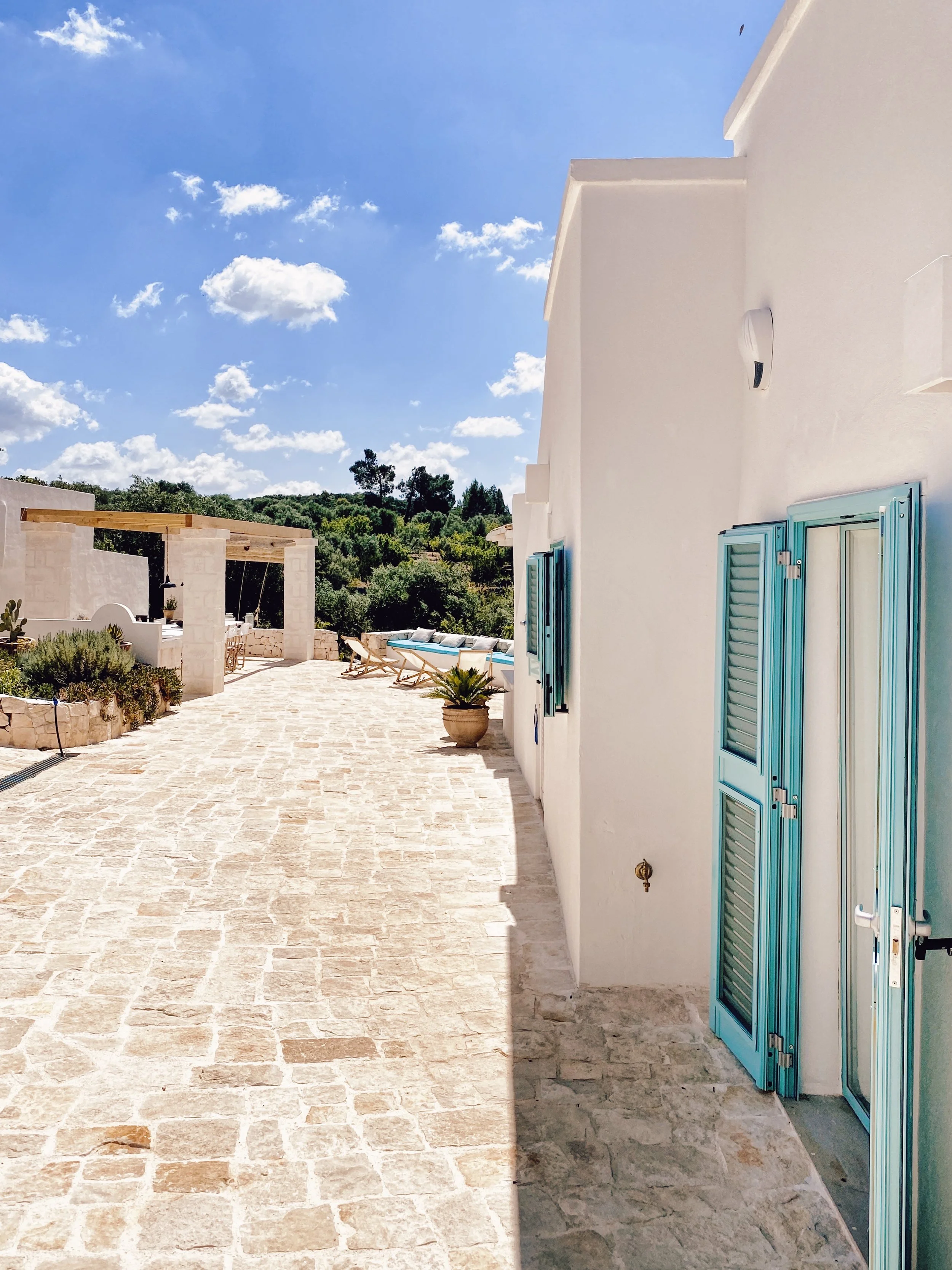 Cortile con pavimentazione in pietra, pareti bianche e serrande azzurre, con sedie a sdraio e piante, sotto un cielo sereno con nuvole sparse.
